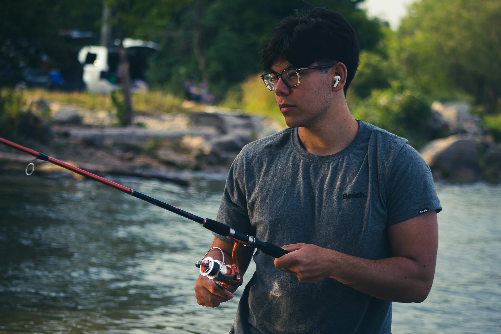 Young man fishing in a river, wearing glasses and wireless earbuds, holding a fishing rod.