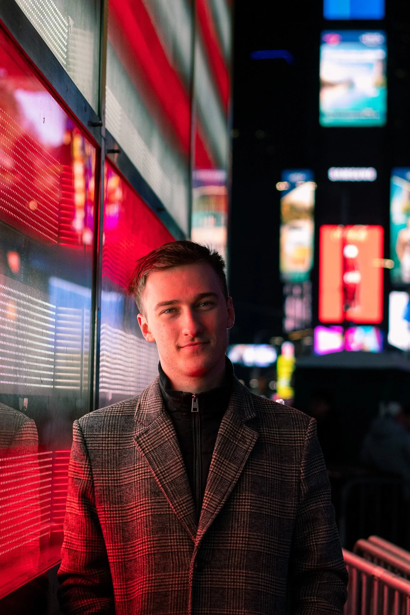 A young man with short hair smiling, standing in front of bright red and multicolored illuminated billboards on a city street at night.