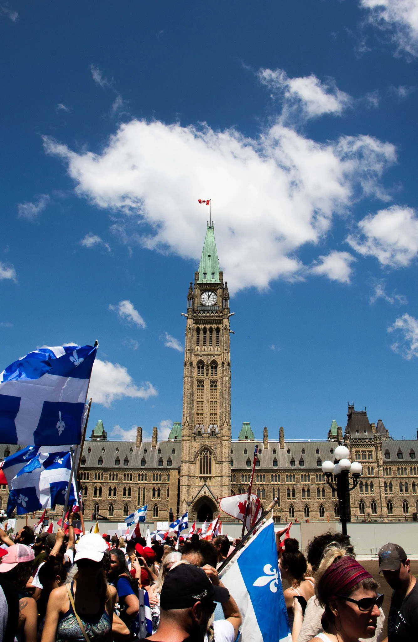 Crowd gathered in front of the Parliament Hill in Ottawa, Canada, with flags and banners, during a sunny day with blue sky and clouds.