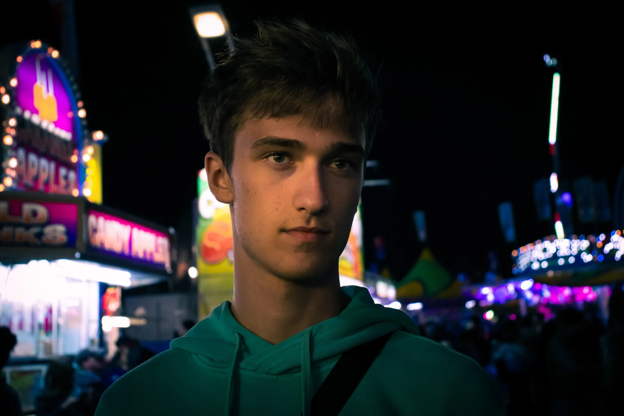 A young man with short dark hair and a green hoodie standing at night in front of bright, colorful neon lights and signs at an amusement park or fair.