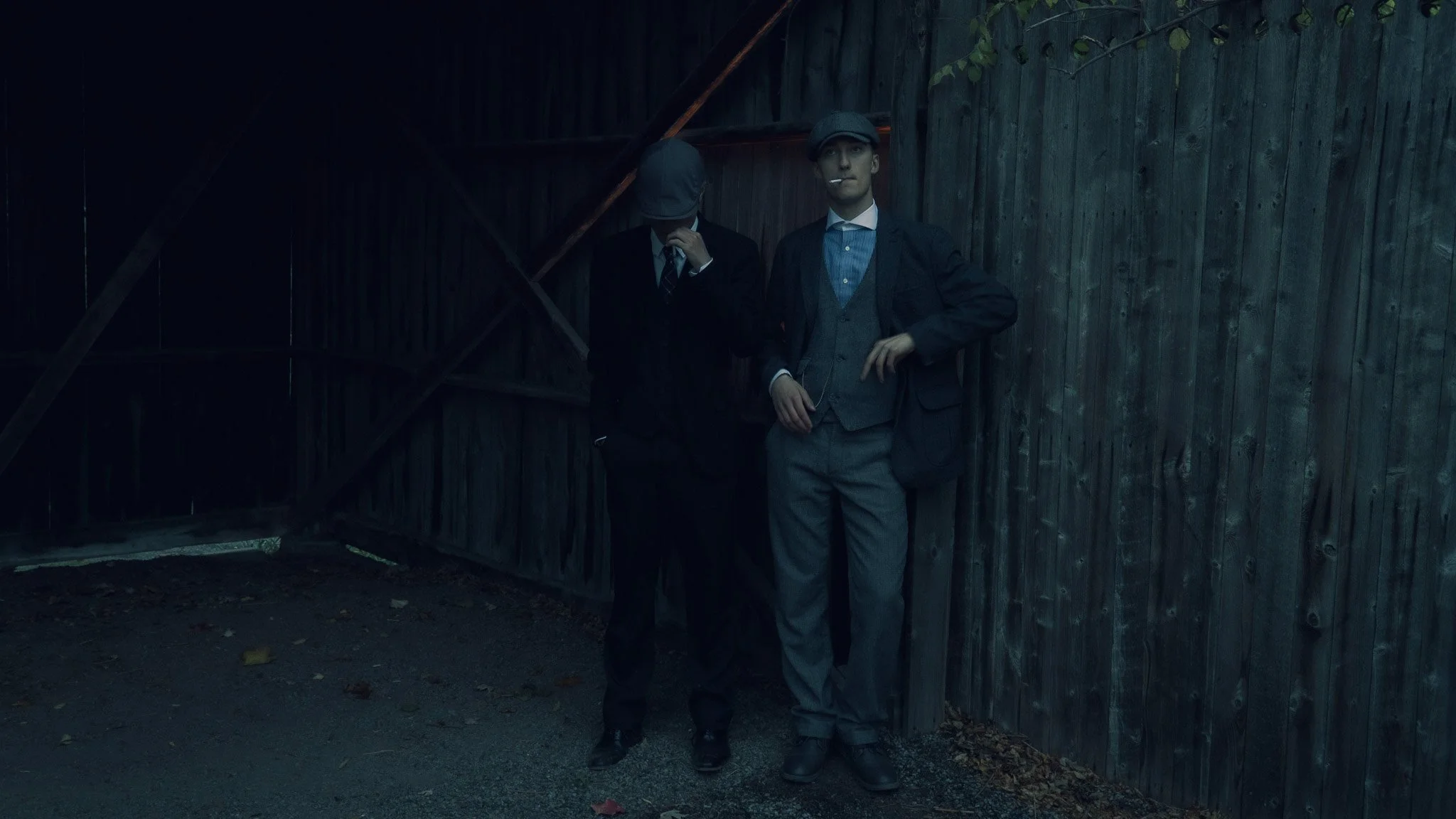 Two men wearing vintage suits and caps standing in front of a wooden fence, one smoking a cigarette, in a dark outdoor setting.