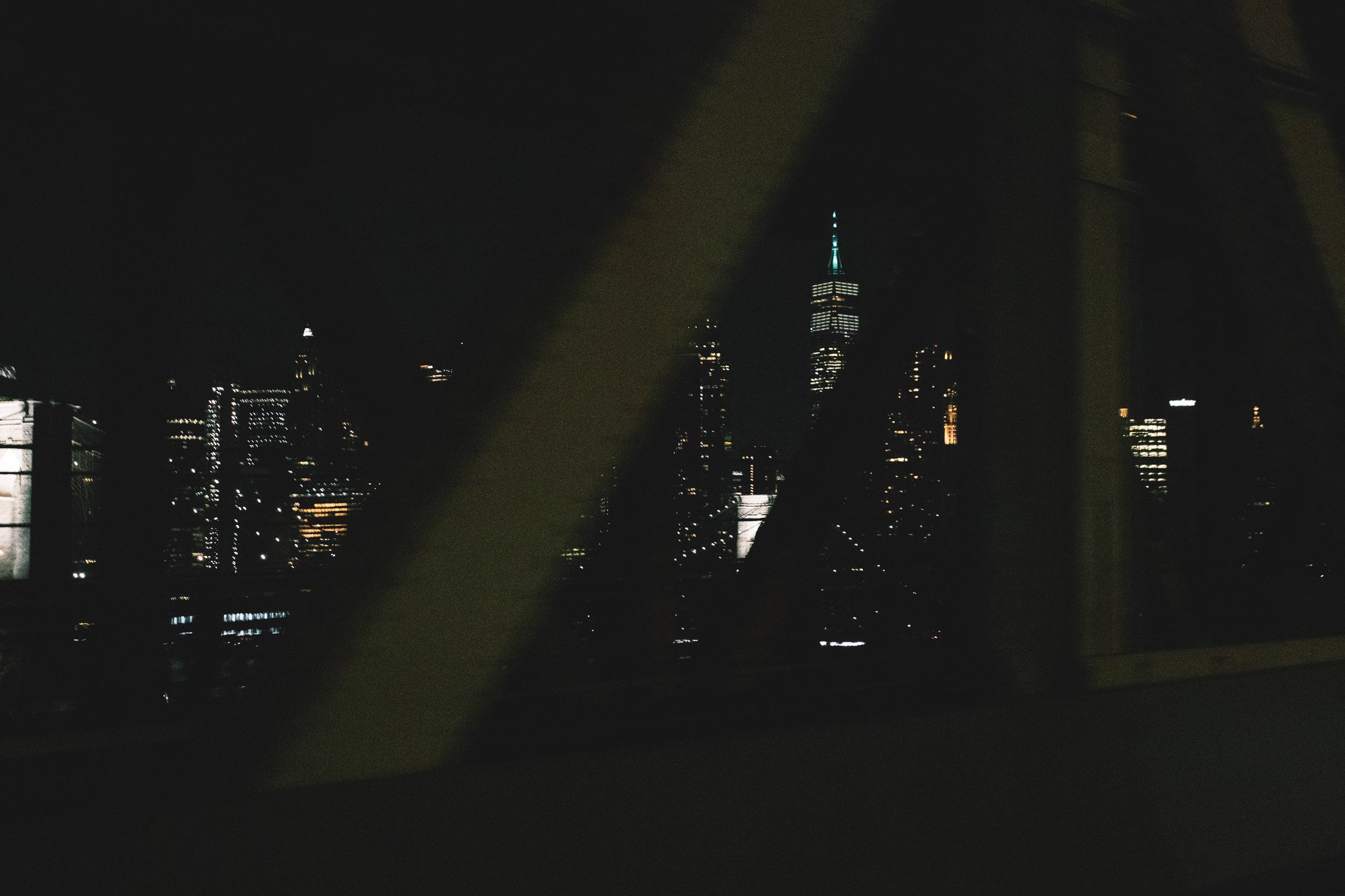 Nighttime cityscape featuring illuminated skyscrapers including the Empire State Building with its spire lit, viewed through a window with dark framing.