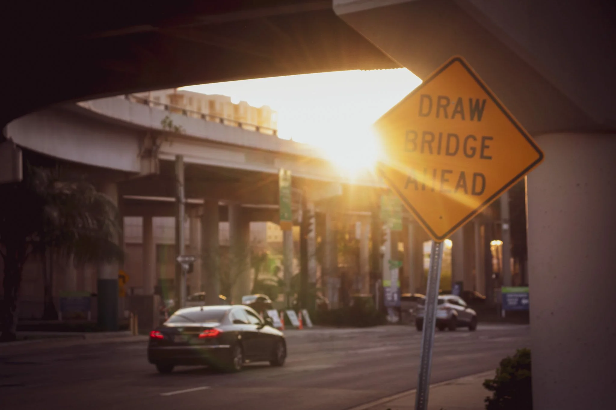A yellow road sign that reads "DRAH BRIDGE AHEAD" is tilted and slightly blurry, with a low sun shining brightly in the background. The scene shows part of a highway with a black car driving underneath the bridge, and an overpass structure overhead.