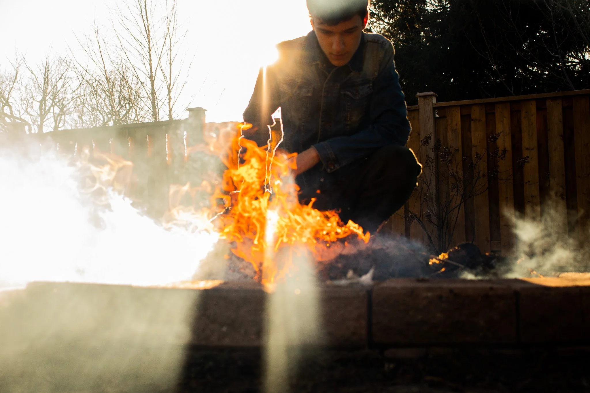 A young man crouches near a fire outdoors during sunset, with a wooden fence and leafless trees in the background.
