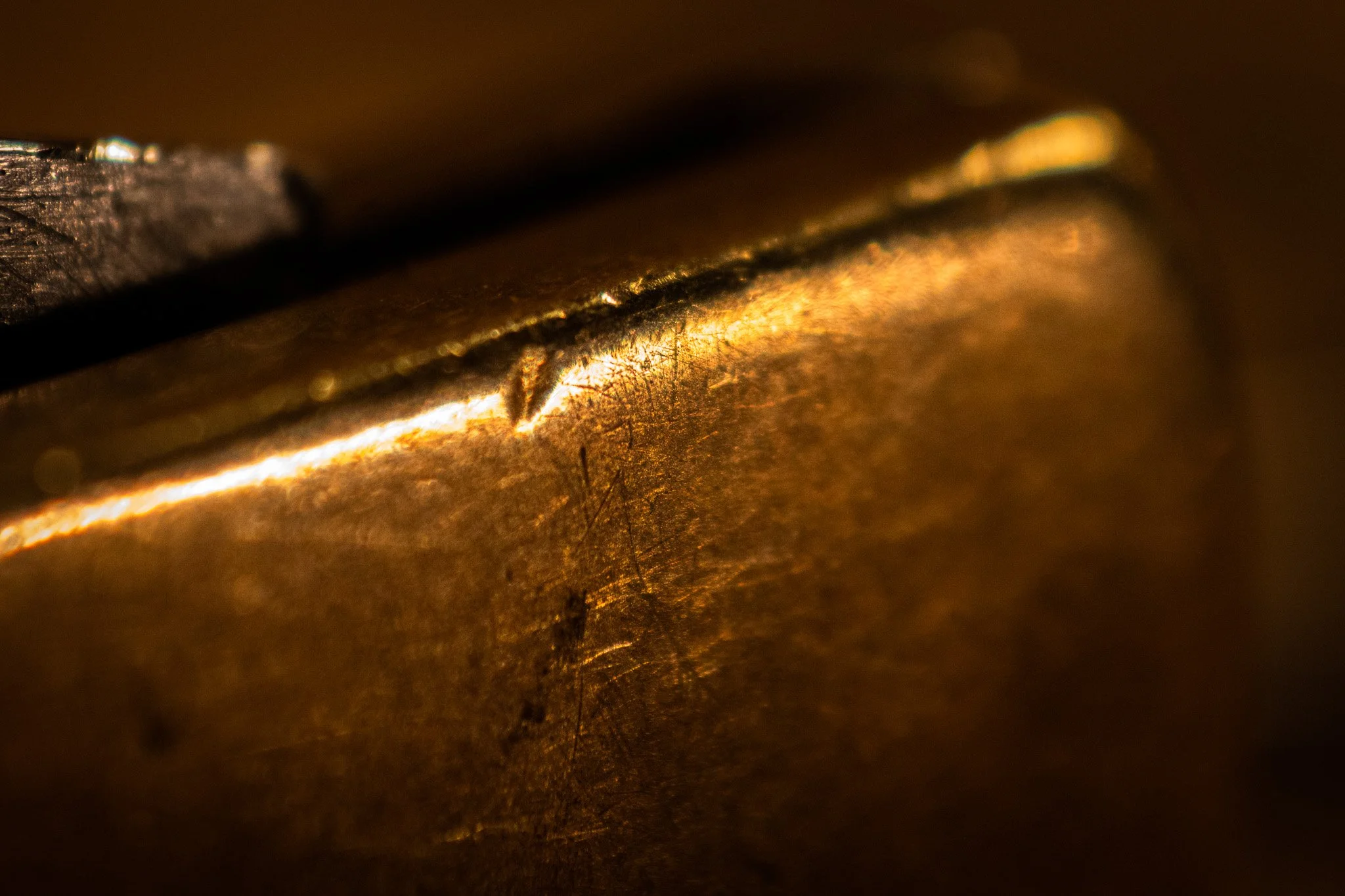 Close-up macro shot of a metallic object with a textured surface, reflecting warm golden and bronze hues, with some darkened areas and scratches.