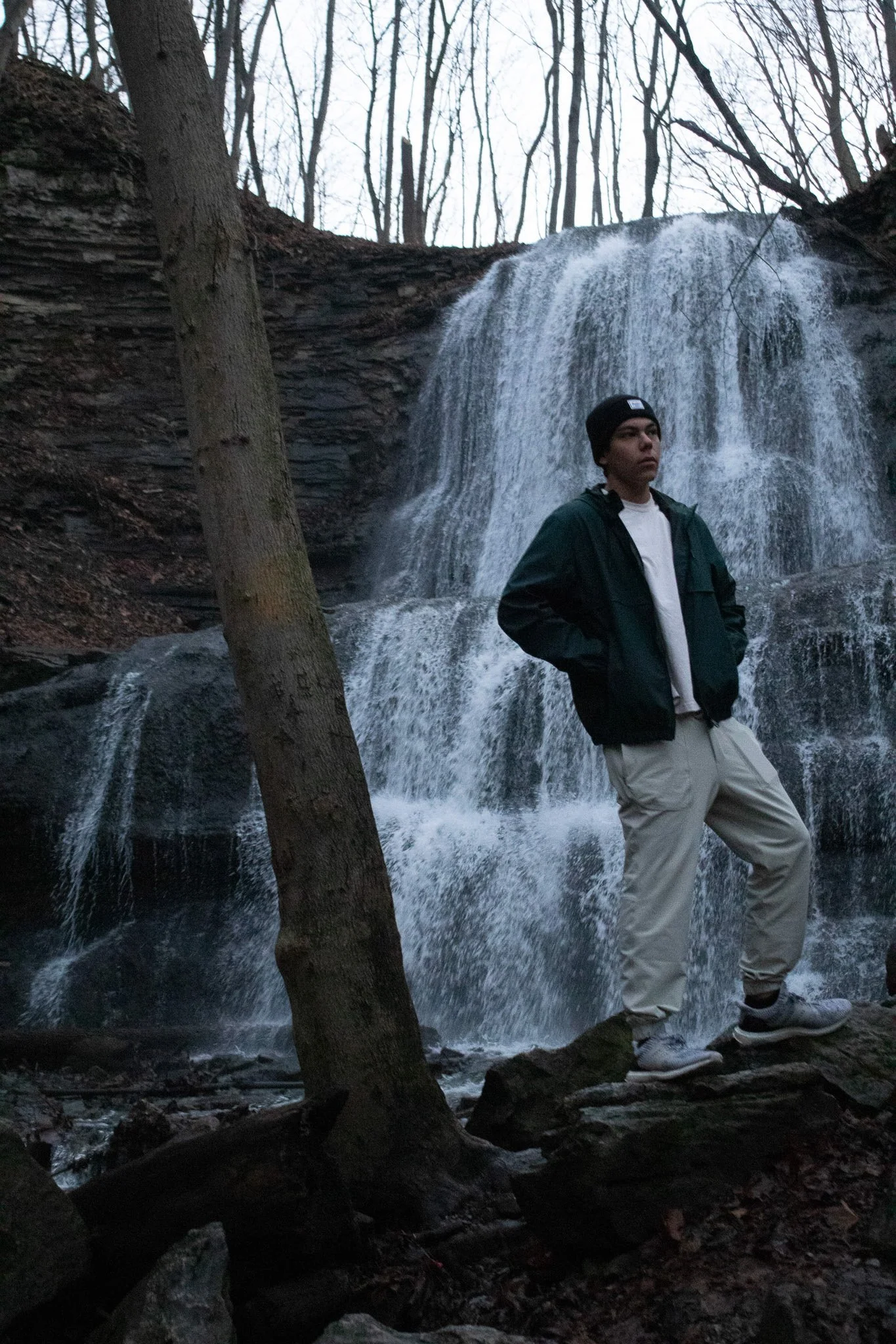 A young man with dark hair, wearing a black beanie, green jacket, white shirt, and light-colored pants, stands by a waterfall in a wooded area with leafless trees.
