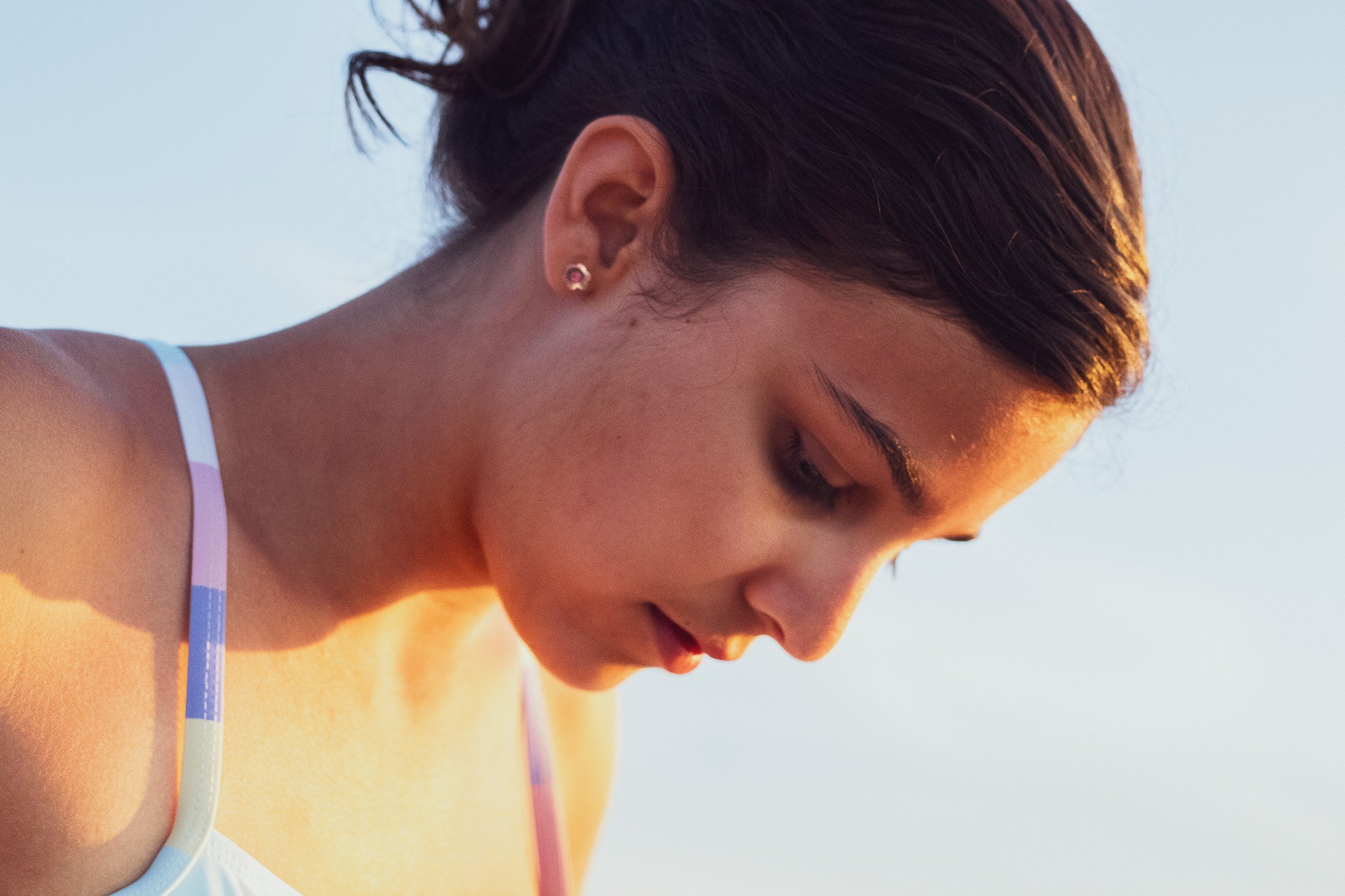 Close-up of a woman with short brown hair wearing a white and pastel-colored swimsuit, looking down with a gentle smile, sunset lighting in the background.