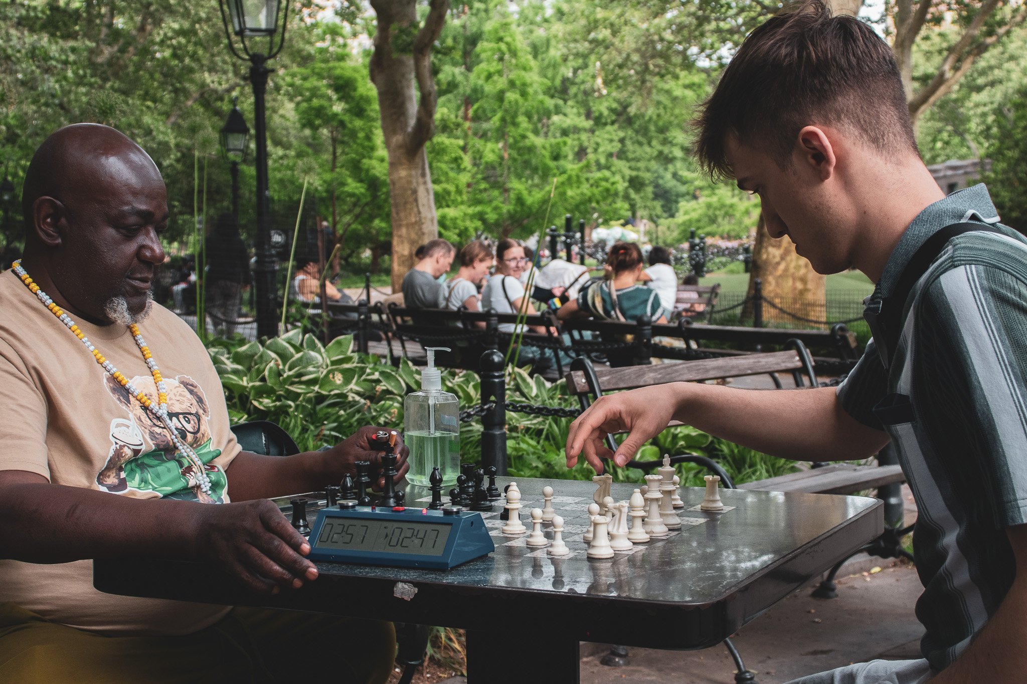 Two men playing chess in a park with green trees and benches in the background. One man is older, bald with a goatee, wearing a beige t-shirt and beaded necklace. The other man is younger with short hair, wearing a striped polo shirt. There is a ches