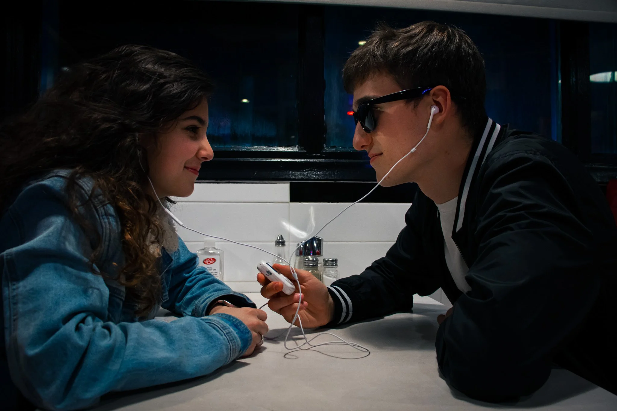 A young woman and a young man are sitting at a table in a dark room facing each other, sharing earbuds, with the woman smiling and the man wearing sunglasses, holding a device. There are salt and pepper shakers and a hand sanitizer bottle on the tabl