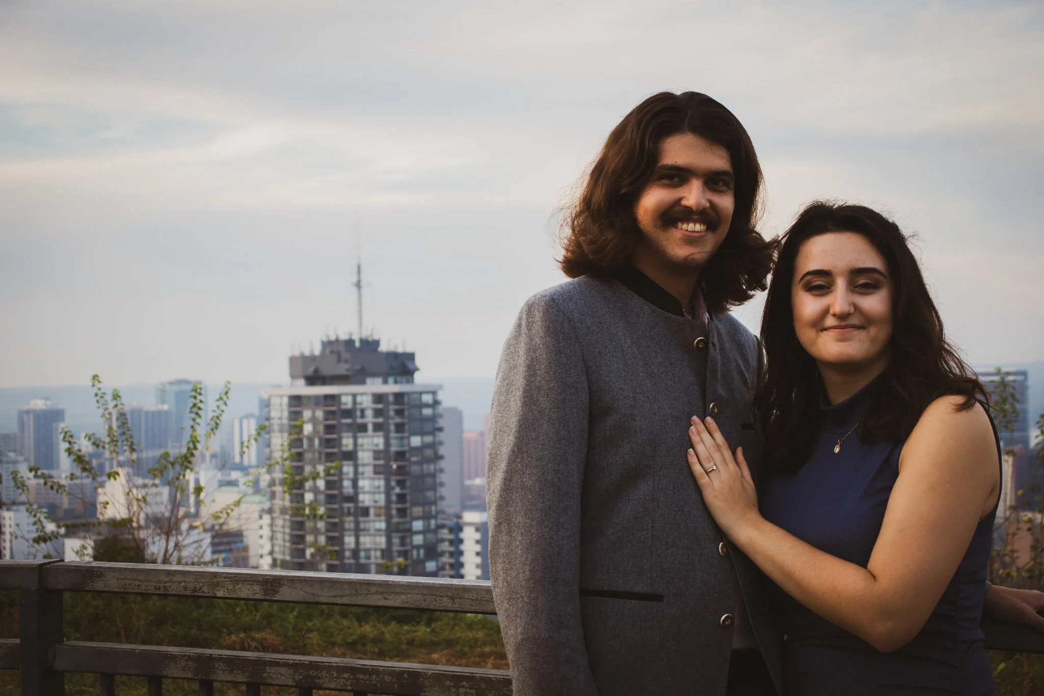 A man and woman standing close together on a balcony with a city skyline in the background, smiling at the camera.