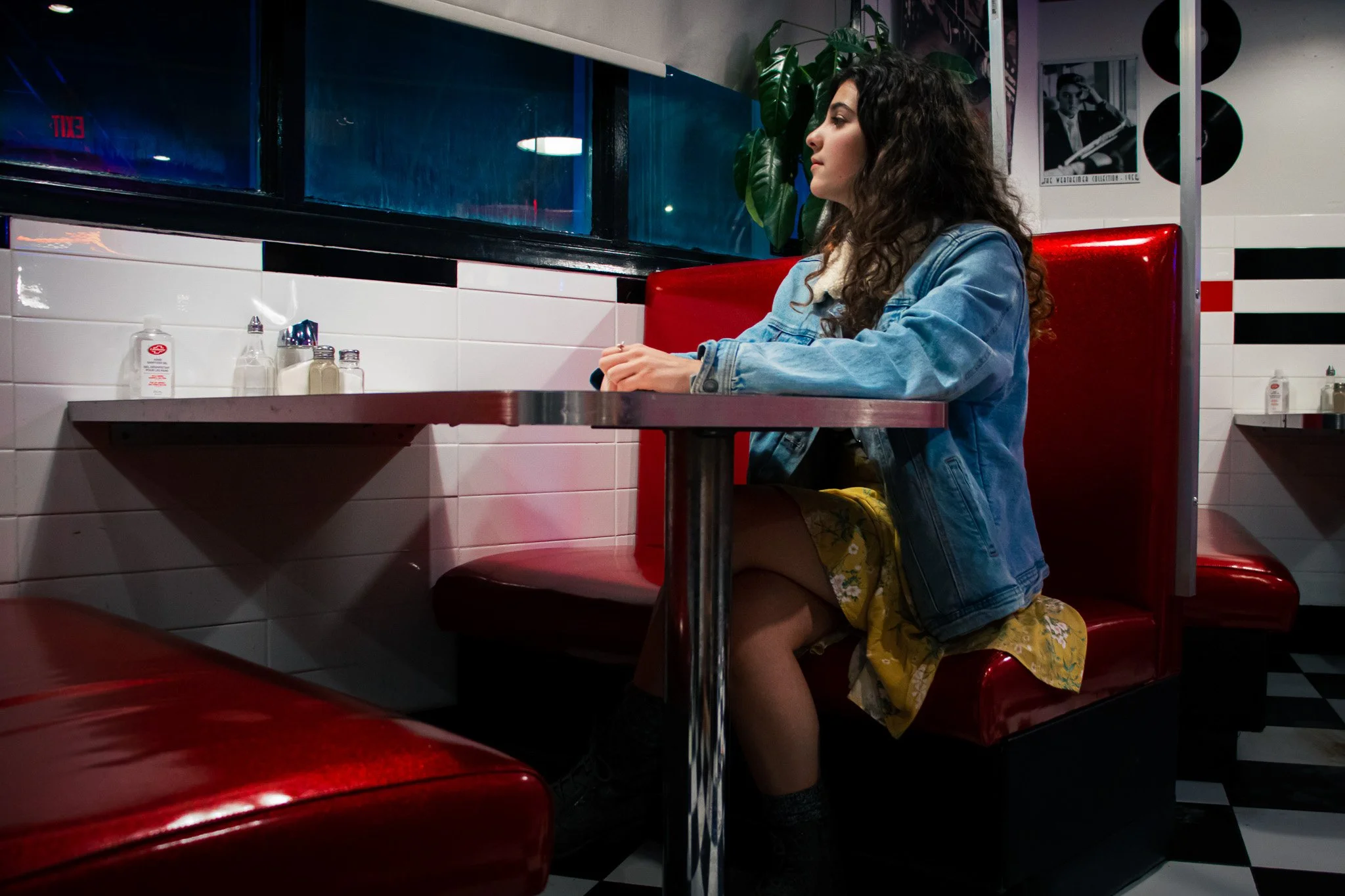 A woman with dark curly hair sitting in a red booth at a retro-style diner, wearing a denim jacket and yellow floral dress, facing the window.