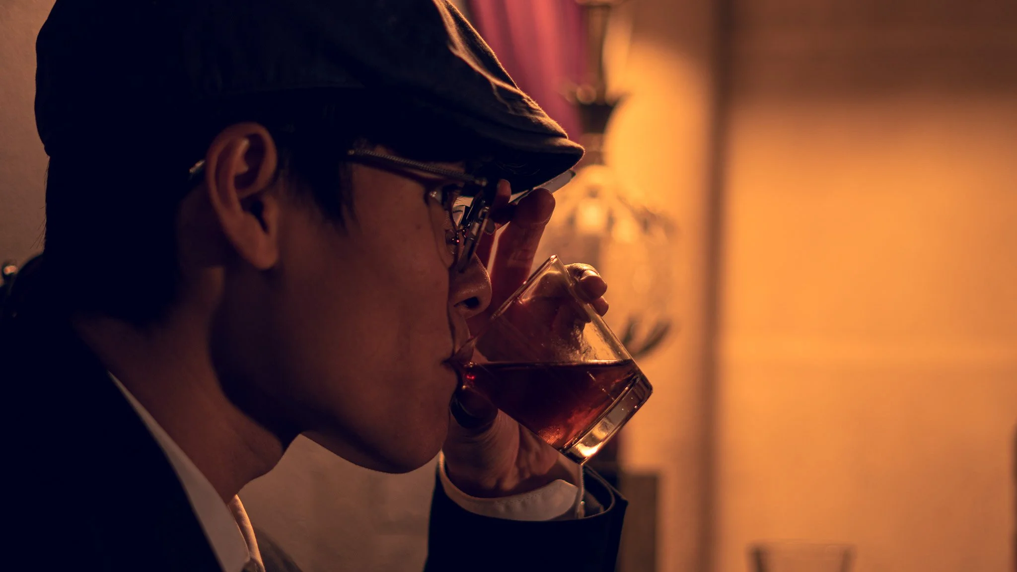 Person wearing glasses and a hat drinking a glass of red wine in a dimly lit room.
