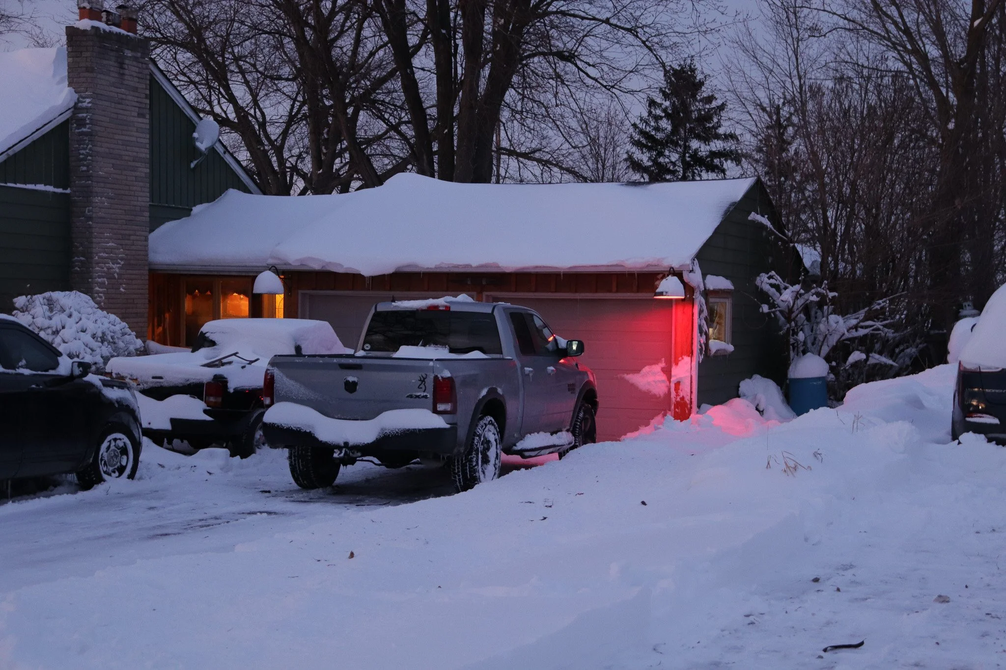 Snow-covered driveway with parked cars, a house with a lit window and snow on the roof, and trees in the background at dusk.