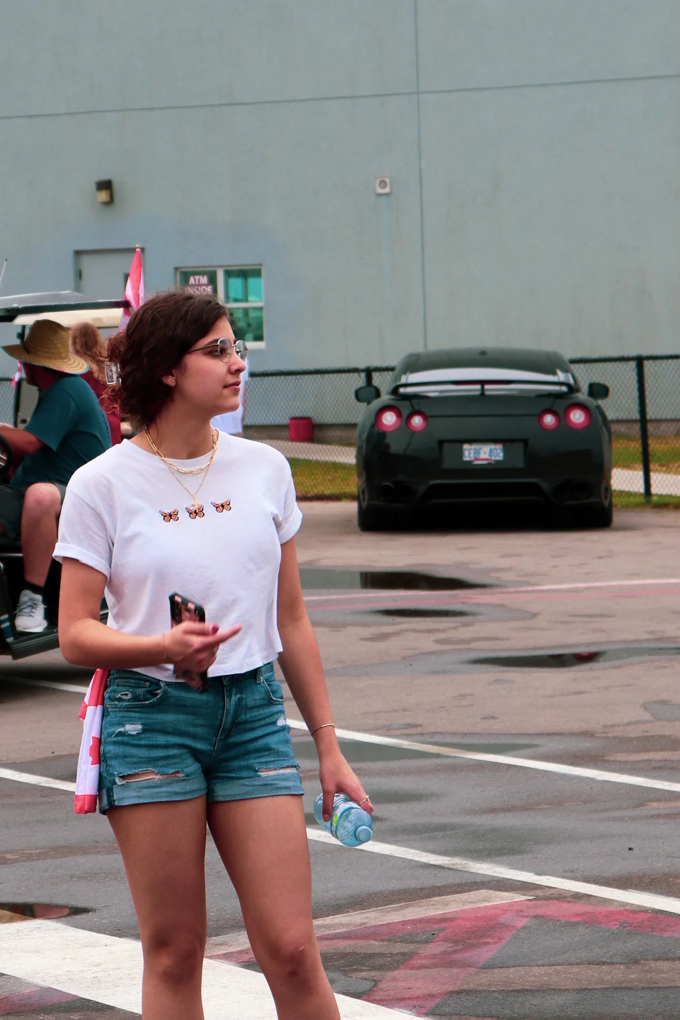 A young woman with short brown hair, sunglasses, a white t-shirt decorated with butterfly pins, and denim shorts walking in a parking lot, holding a water bottle, with a background of a gray building, a black sports car, and other people.