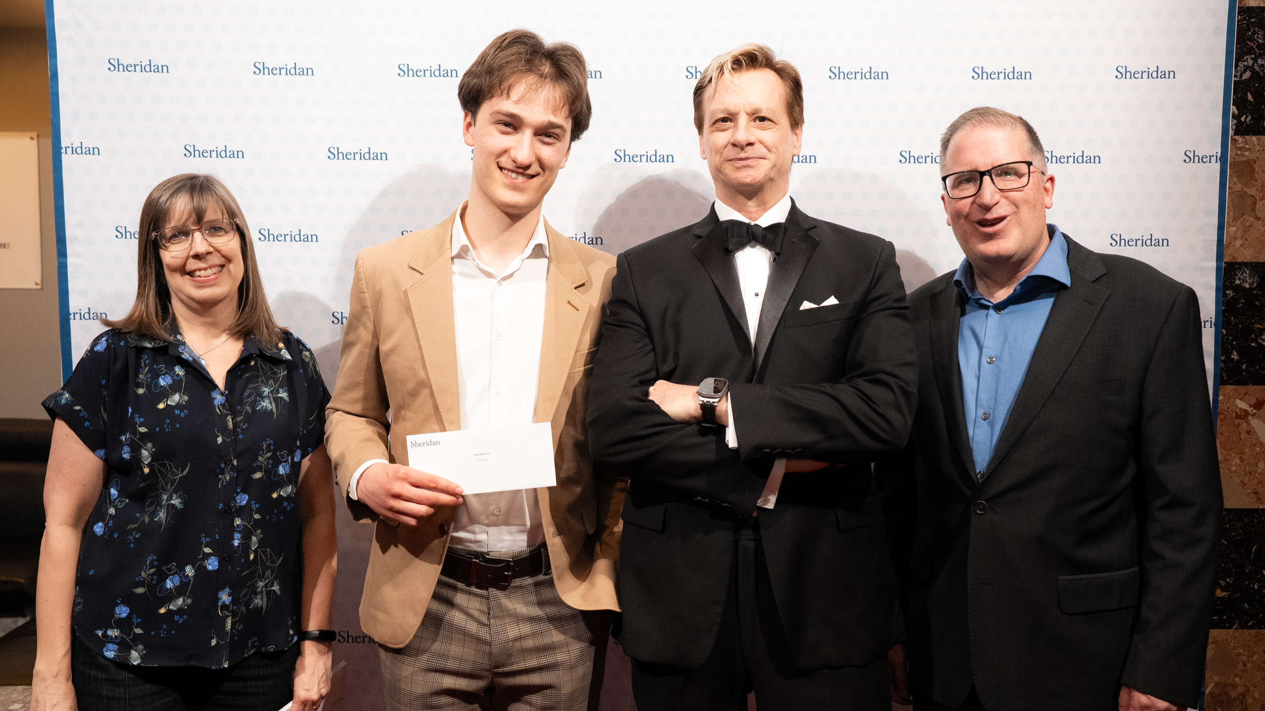 Group of four people at a Sheridan event, smiling and posing for a photo, with a Sheridan backdrop behind them.