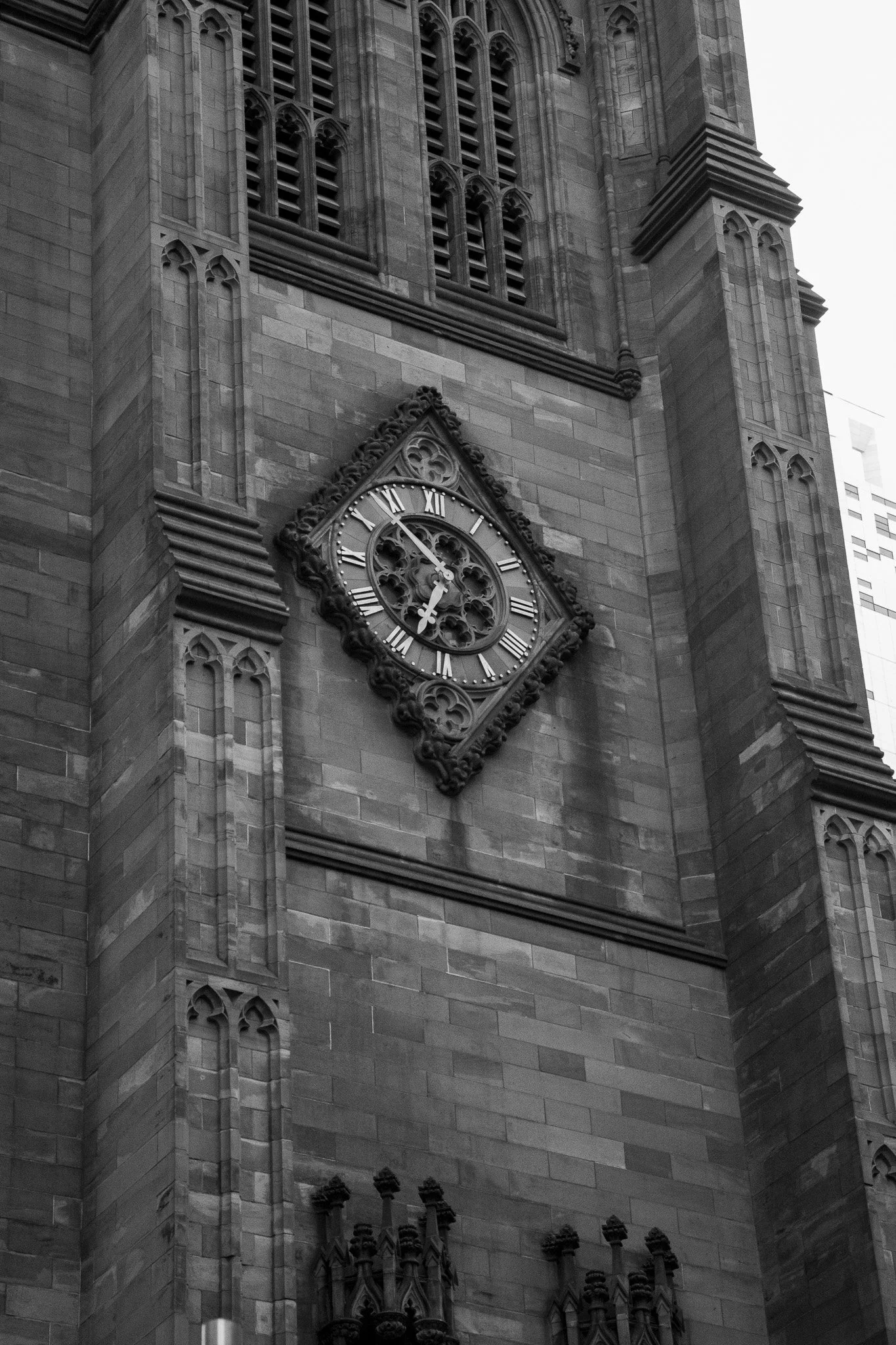 A black and white photograph of a historic stone church or cathedral, featuring a clock with Roman numerals on its tower and gothic architectural details such as arched windows and decorative stonework.