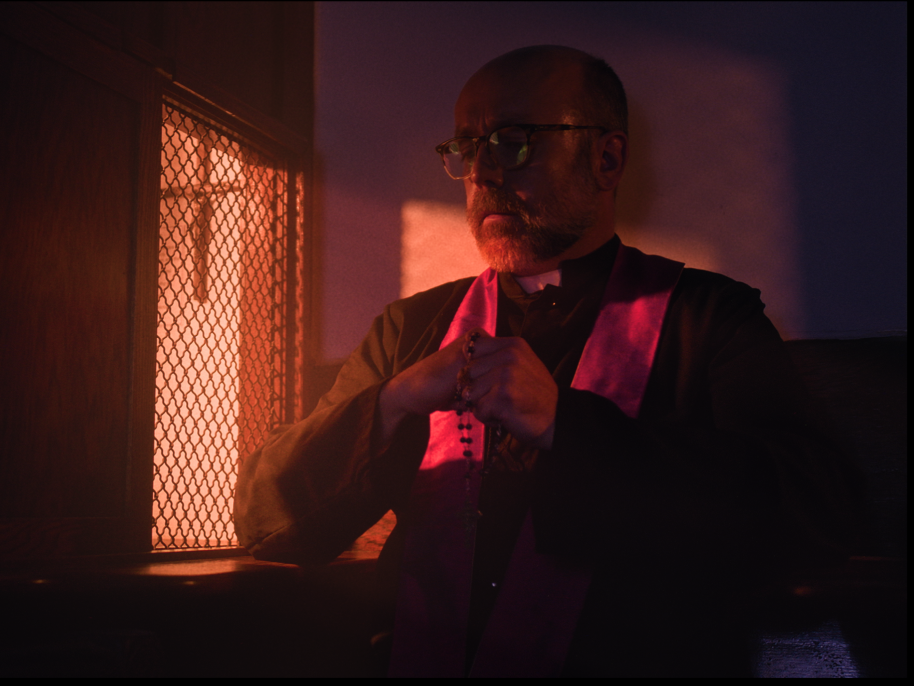 A man with glasses and a beard is sitting in a dimly lit room, wearing a black shirt and a pink stole. He is holding prayer beads with both hands, in front of a window with an orange-red glow.
