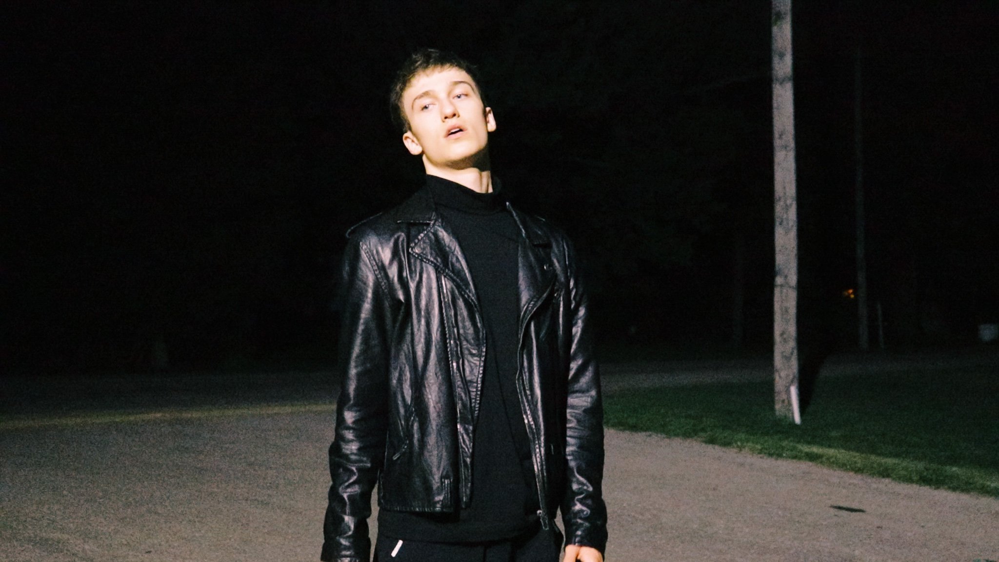 Young man standing outdoors at night wearing a black leather jacket and black turtleneck, with a dark background and a utility pole nearby.