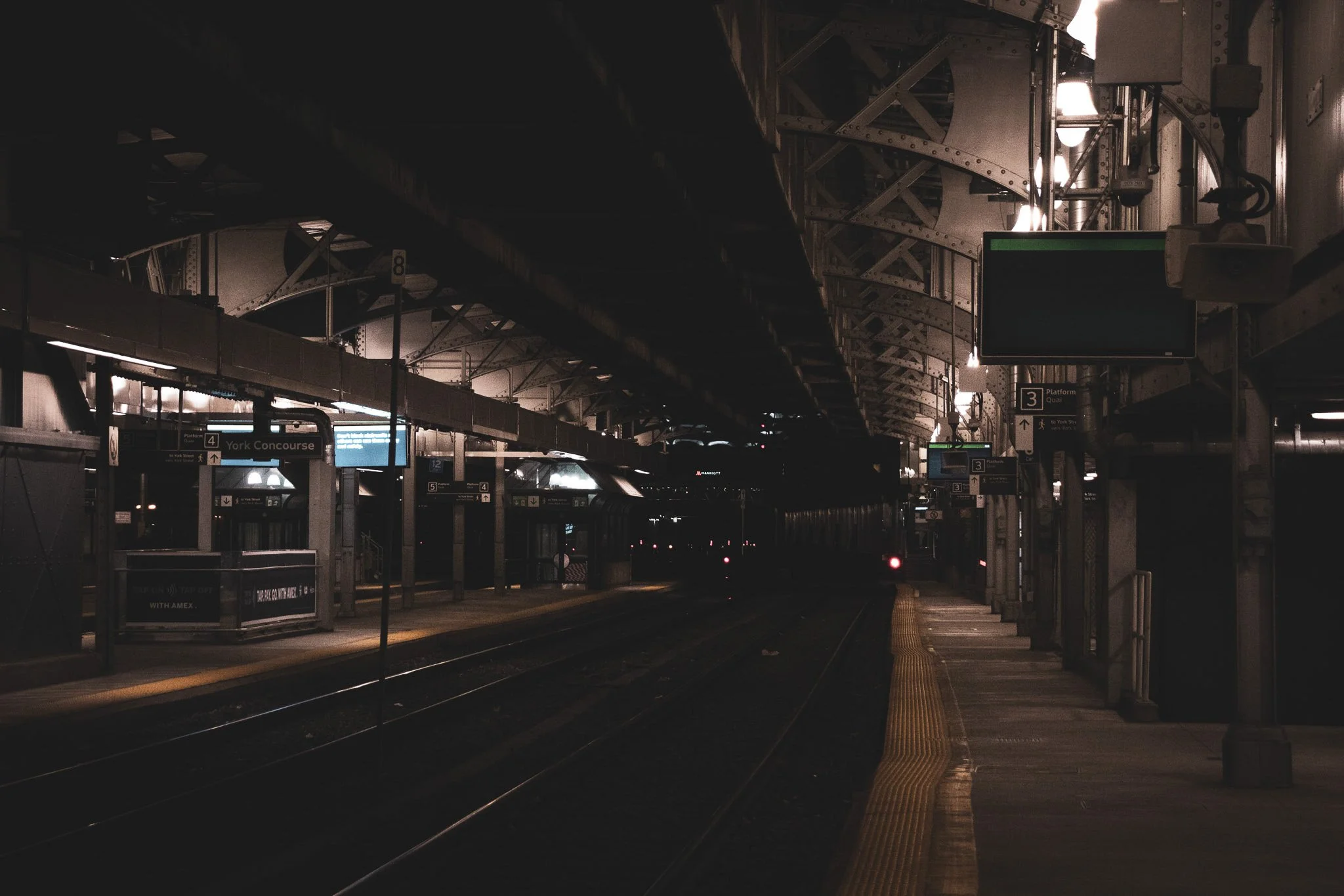 Empty subway platform at night with overhead lighting, digital signs, and tracks extending into the distance.