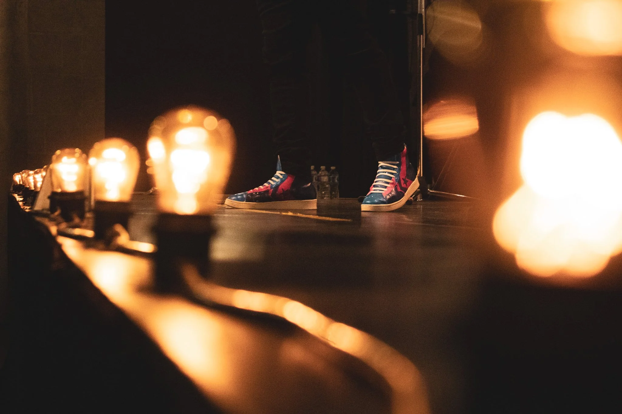 Dimly lit scene with a person's legs wearing colorful, patterned high-top sneakers, standing on a dark surface surrounded by lit candles.