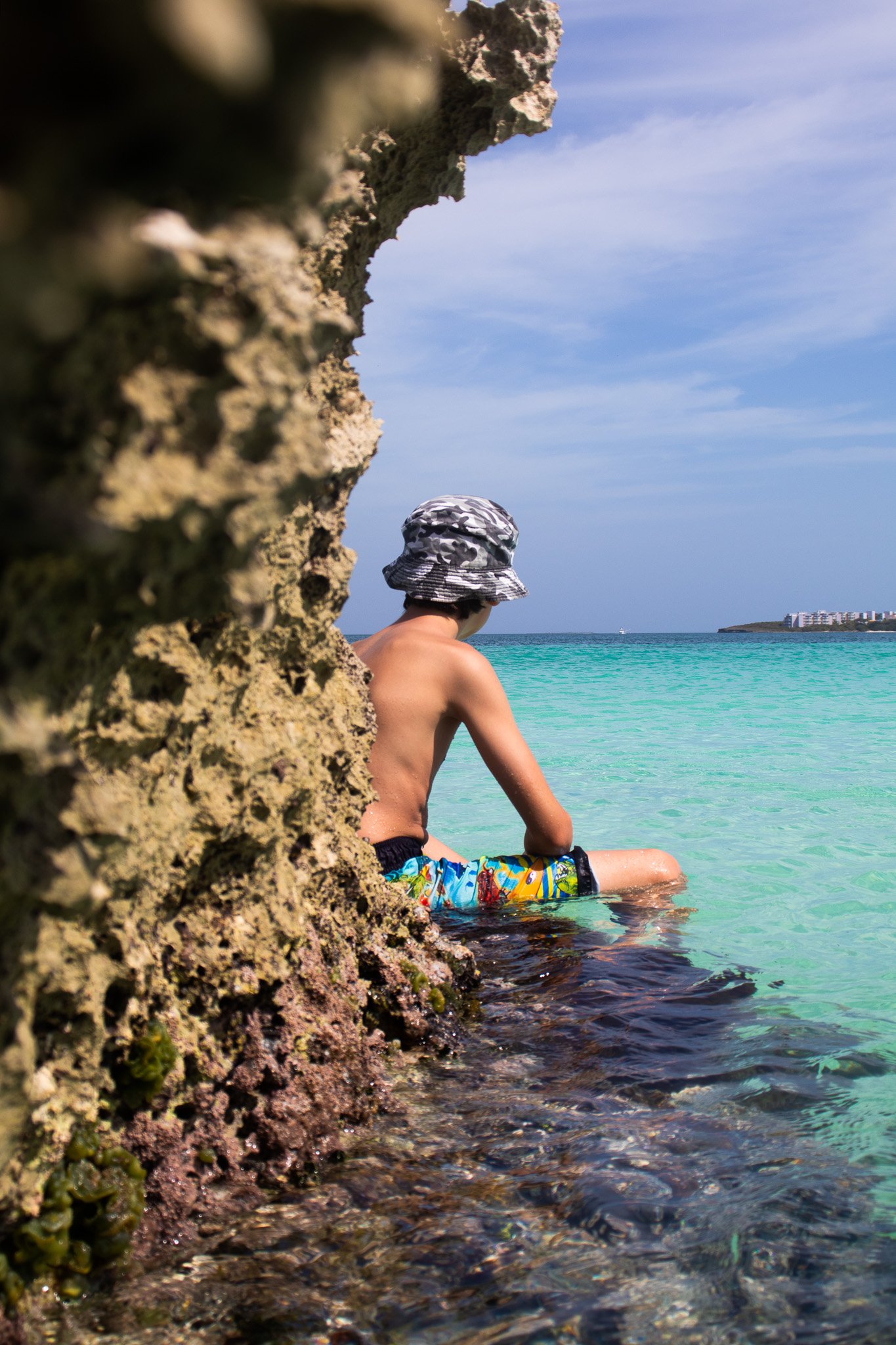 A shirtless man wearing a camo bucket hat and colorful swim trunks sitting by the turquoise sea, partially hidden behind a rugged rock formation with green moss or algae, overlooking a distant island with buildings.