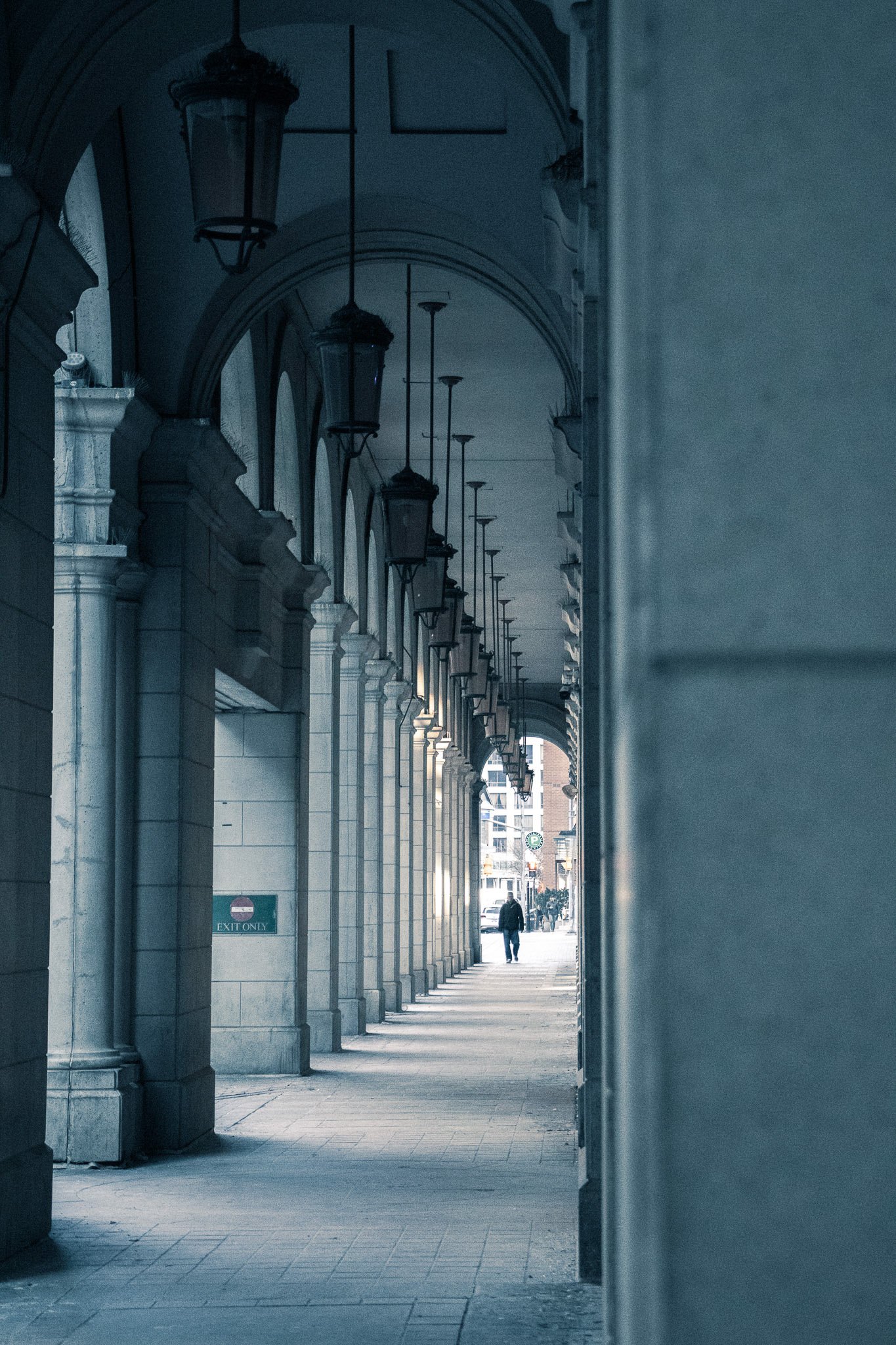 View through an arched colonnade with hanging lamps, leading to a street with a person walking in the distance.