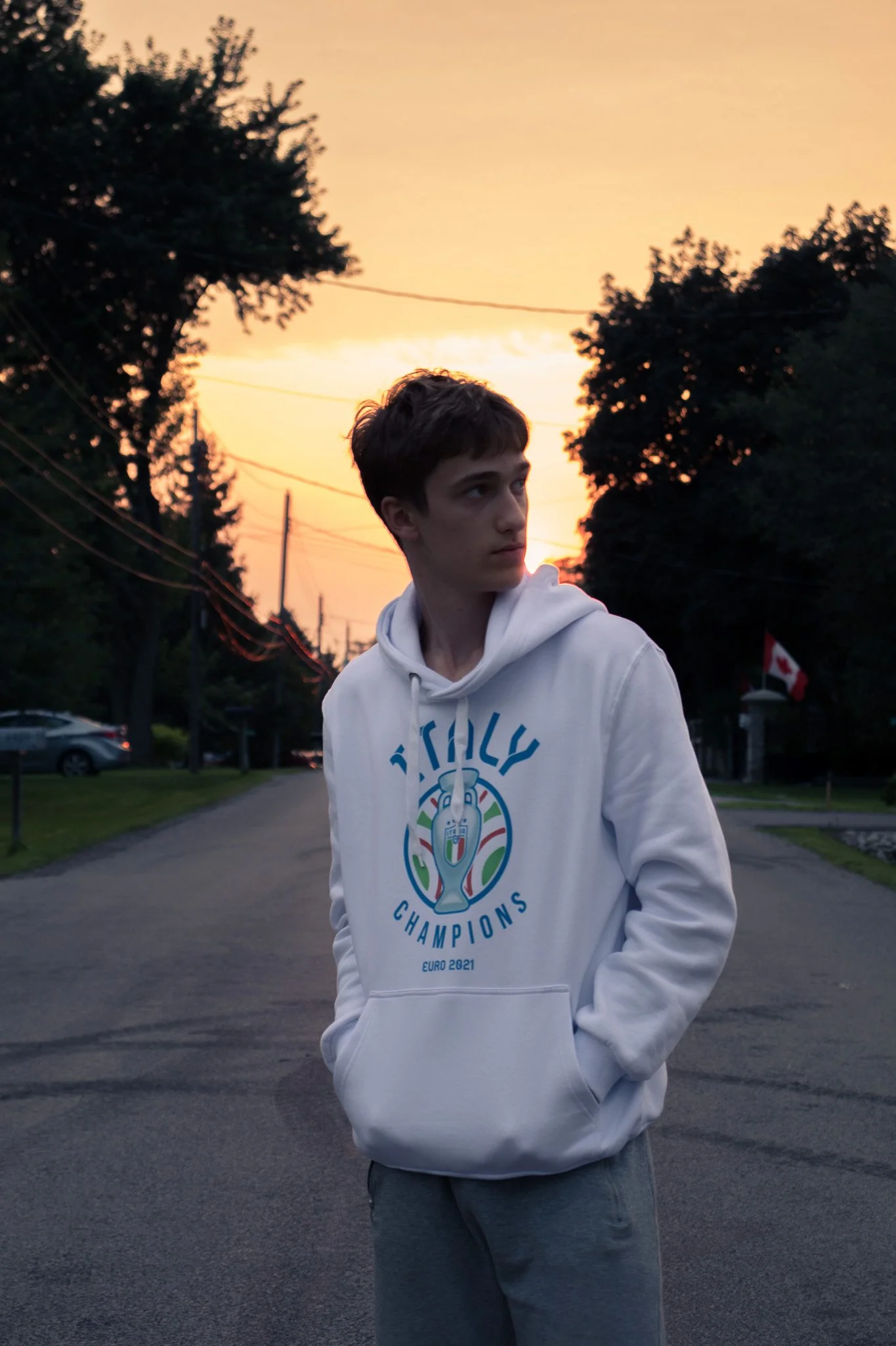 A young man with dark hair and light skin wearing a white hoodie with a Euro 2021 champion logo, standing outdoors at sunset. Trees, power lines, and a Canadian flag in the background.