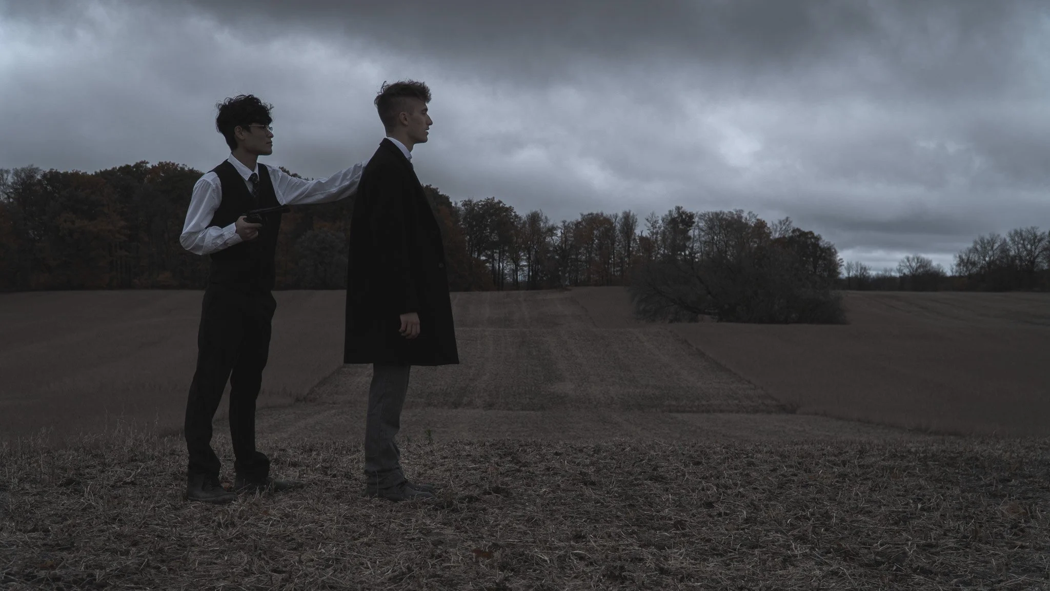 Two young men standing outdoors on a cloudy day, one holding a gun and placing a hand on the other's shoulder, with a forested landscape in the background.
