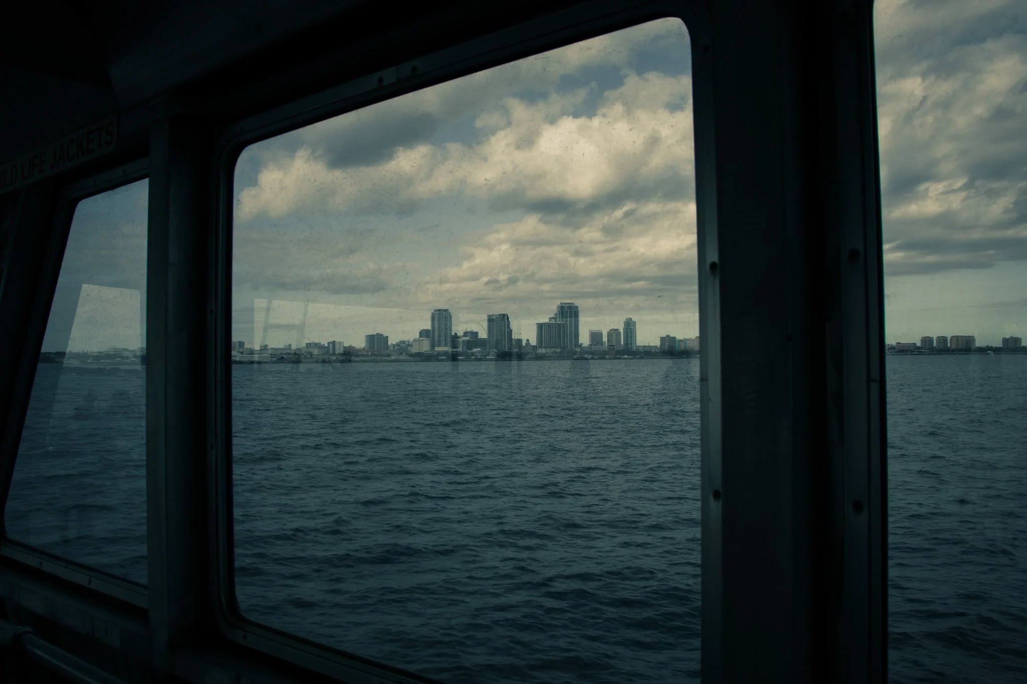 View of a city skyline with tall buildings seen through a window of a boat, water in the foreground, cloudy sky above.