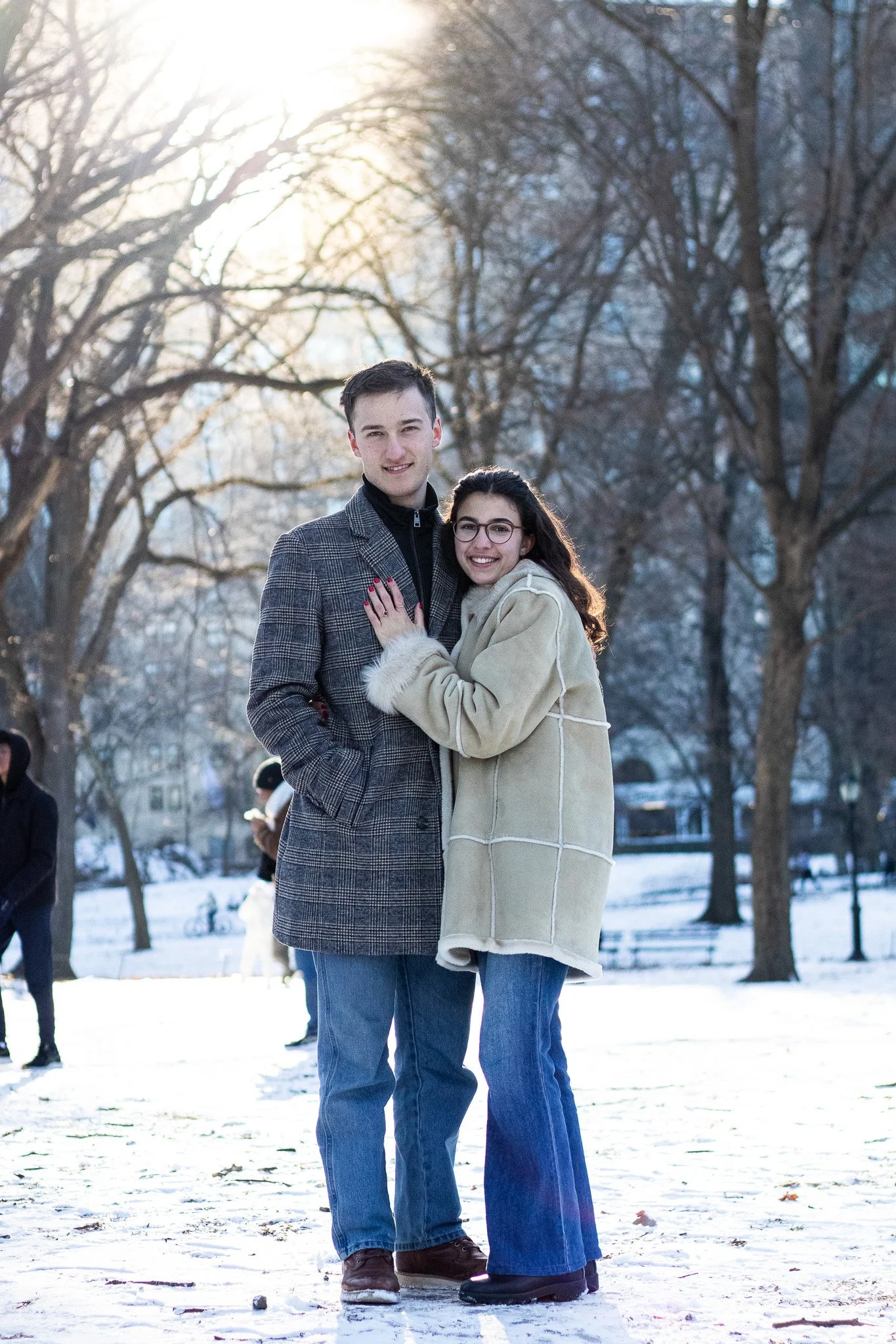 A young couple standing close together outdoors on a snowy day, smiling and embracing, with bare trees and a bright sun in the background.