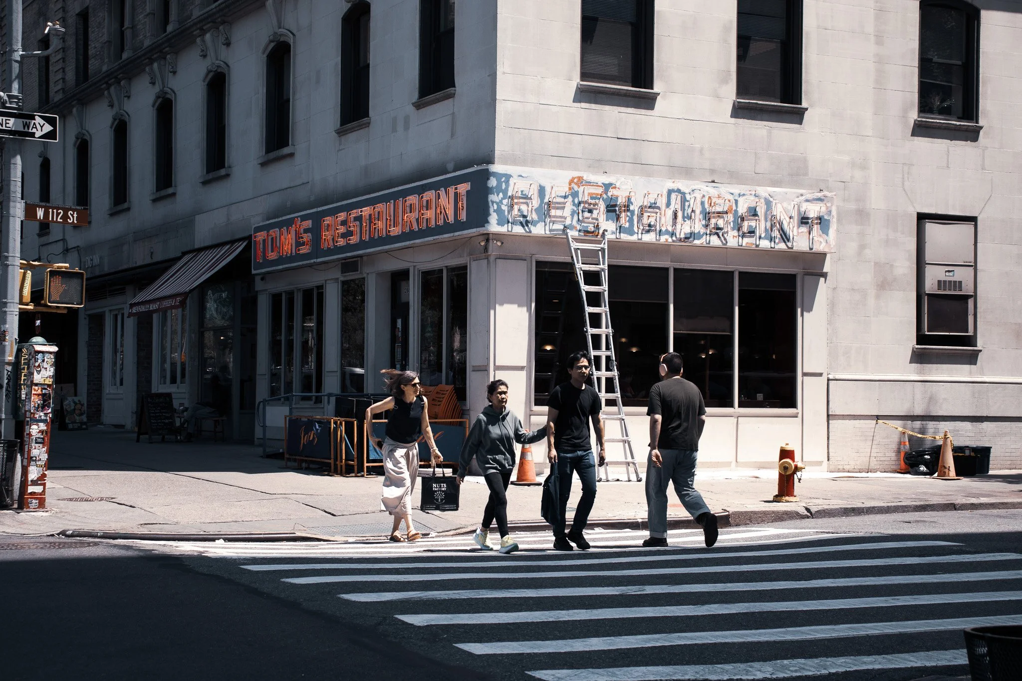 People crossing the street in front of a corner building with a sign for Tom's Restaurant at W 112th Street, Manhattan.