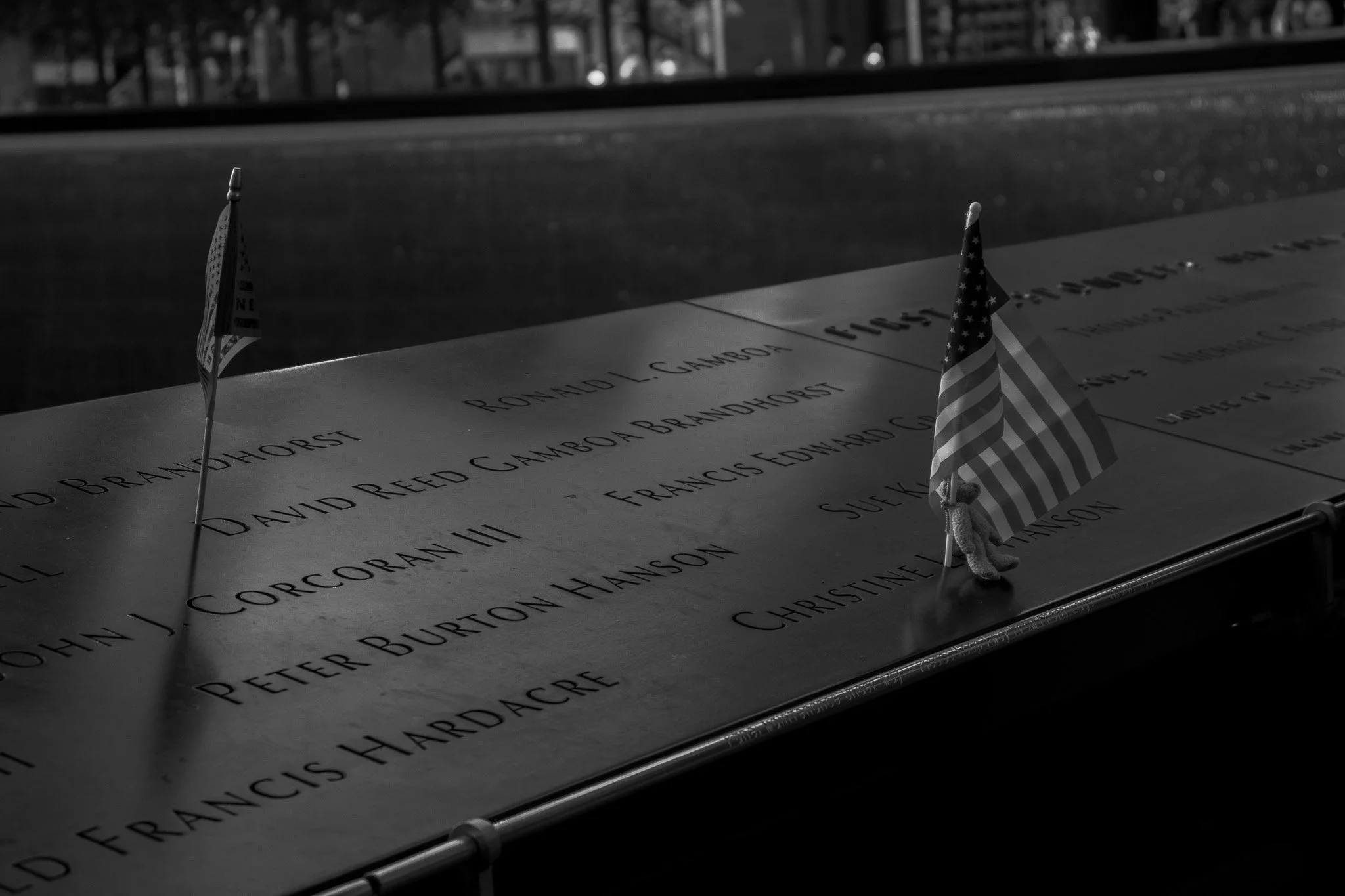 Close-up of the Vietnam Veterans Memorial with engraved names of fallen soldiers. Two small American flags are placed among the names, and a pencil is shown at the bottom right corner.
