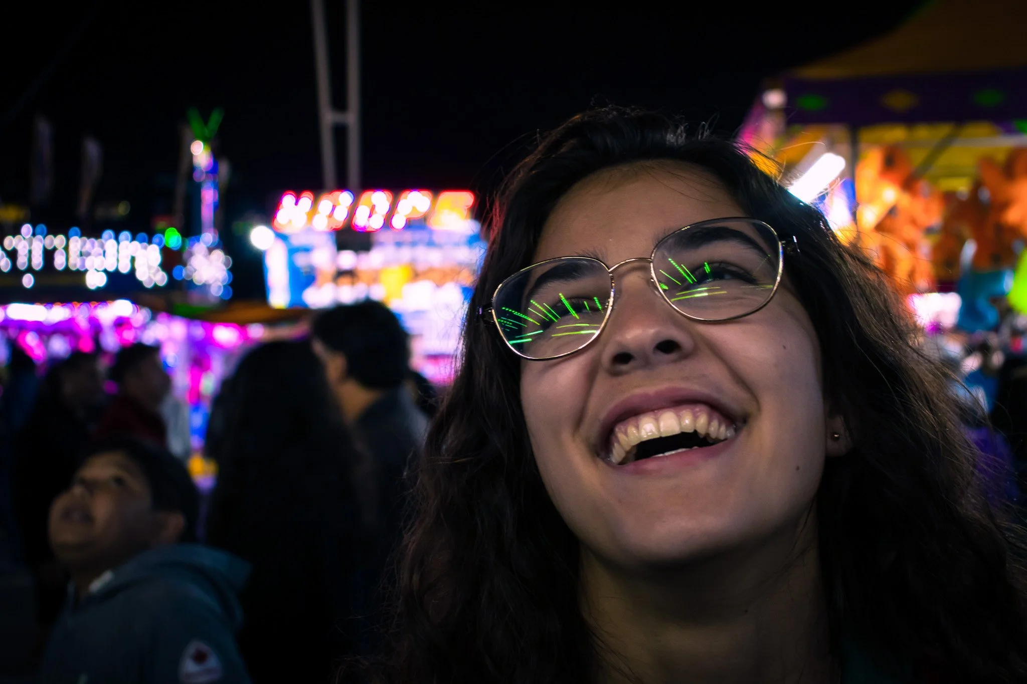 A person with glasses smiling at a nighttime carnival with colorful lights and blurred crowd.