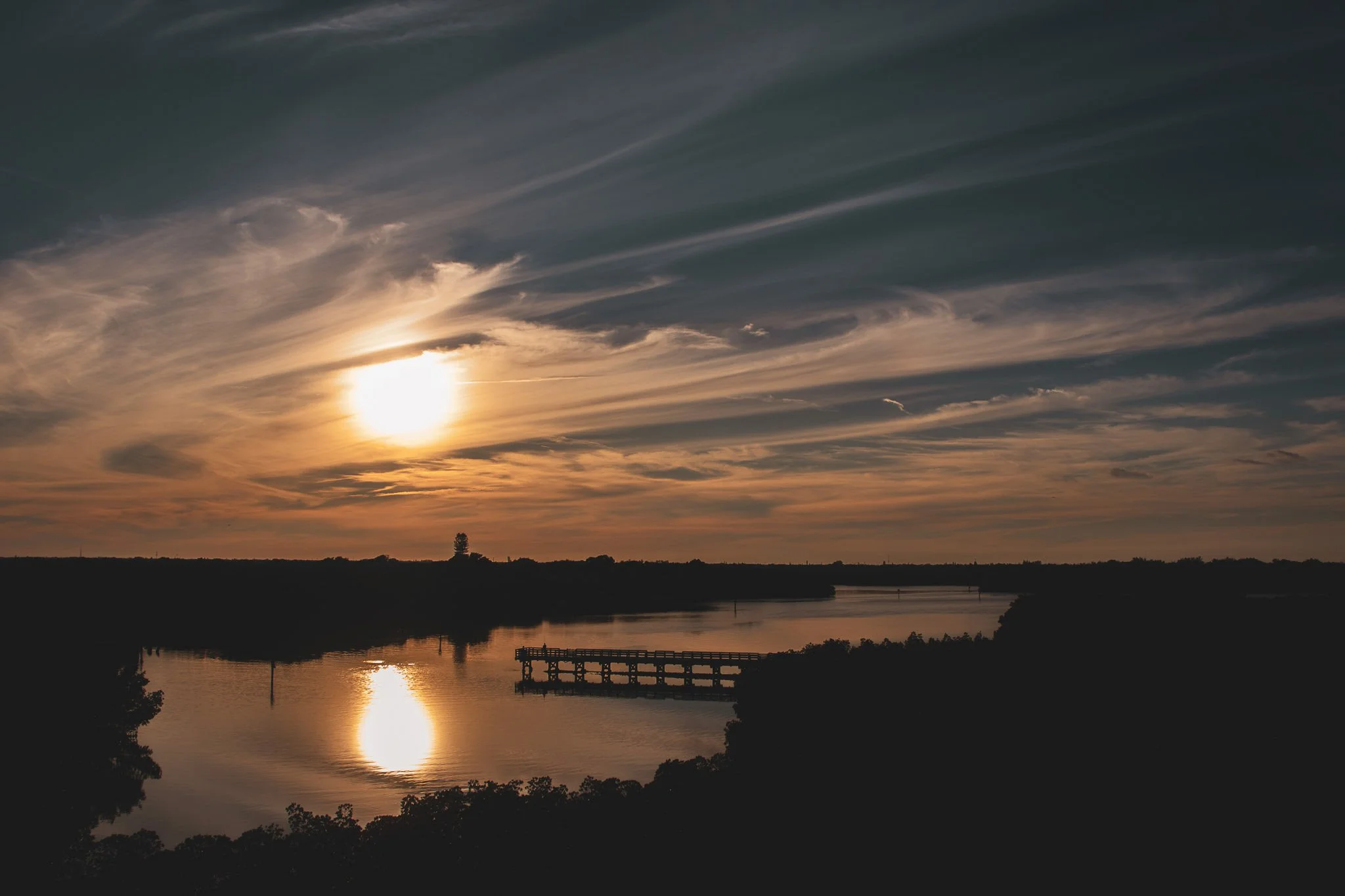 Sunset over a river with reflections, silhouette of trees and a distant pier or dock.