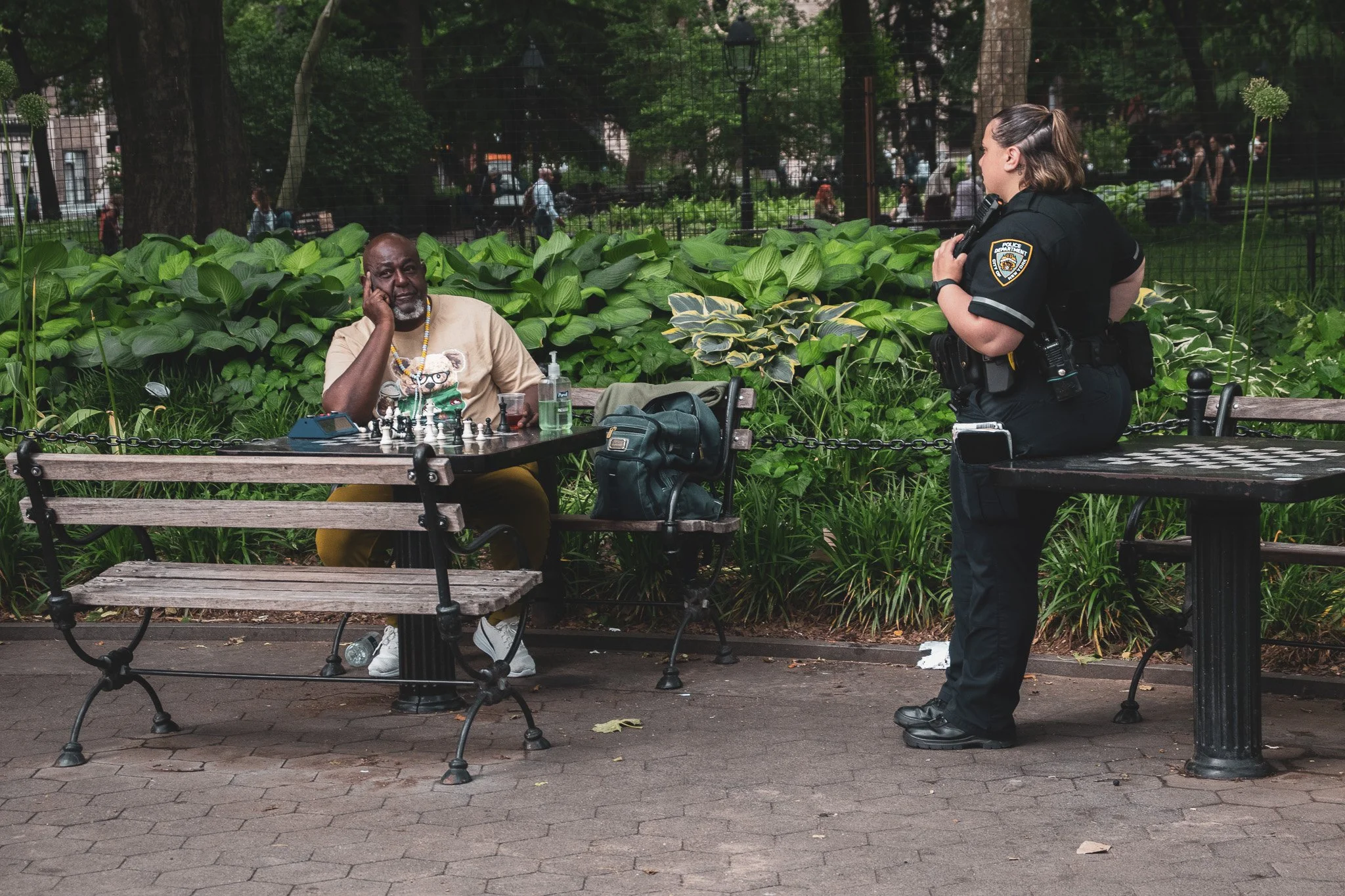 A police officer talks with an elderly man sitting at a park bench while playing chess. The man appears thoughtful, resting his face on his hand. Items like a water bottle, a small electronic device, and a backpack are on the table. The background sh