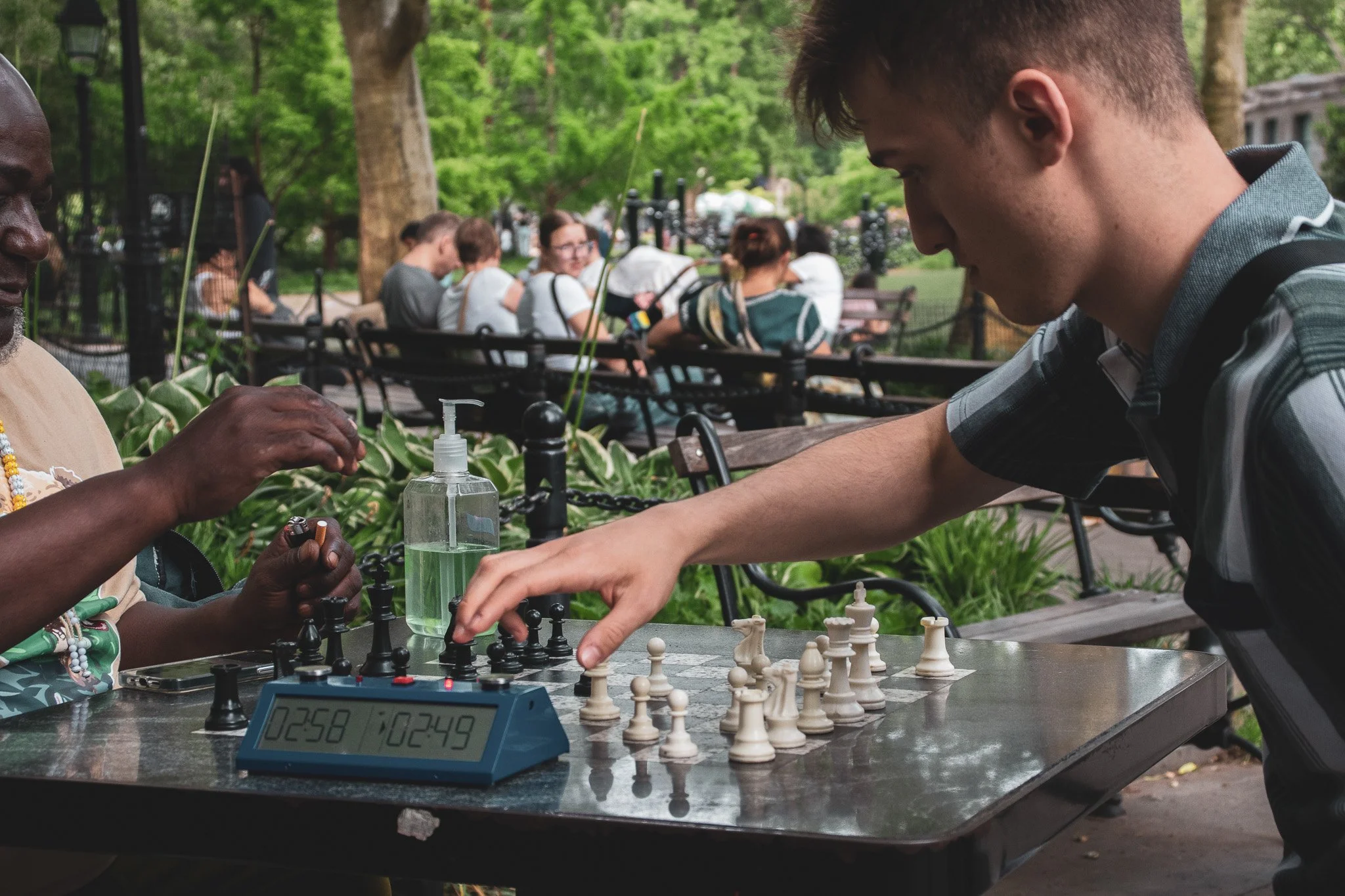 Two people playing chess at an outdoor park table amidst green trees. The person on the left is a woman with dark skin, wearing a beige outfit with beads and a necklace. The person on the right is a man with light skin and short hair, wearing a black