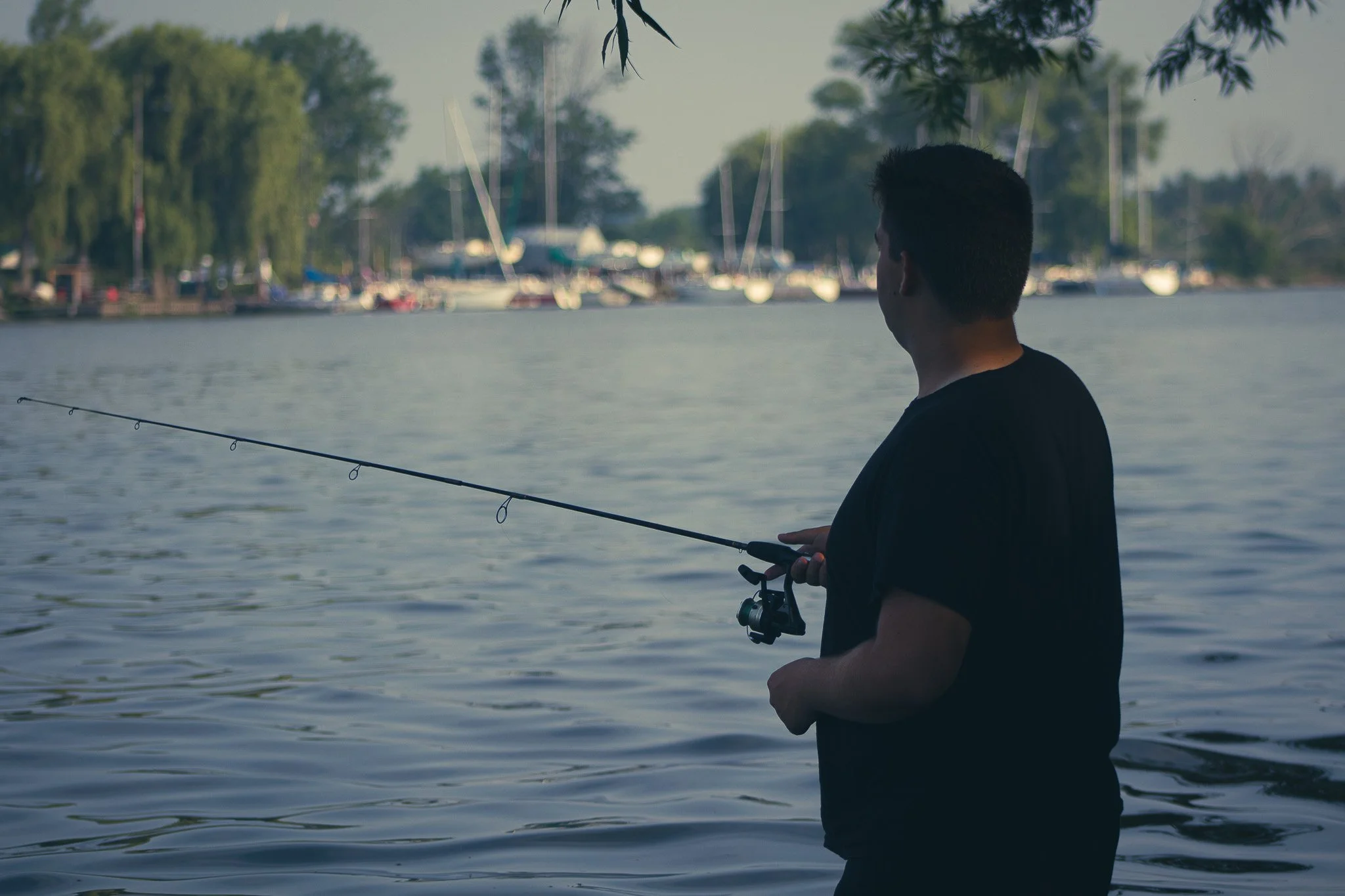A young man is fishing by the water, holding a fishing rod, with boats and trees in the background.