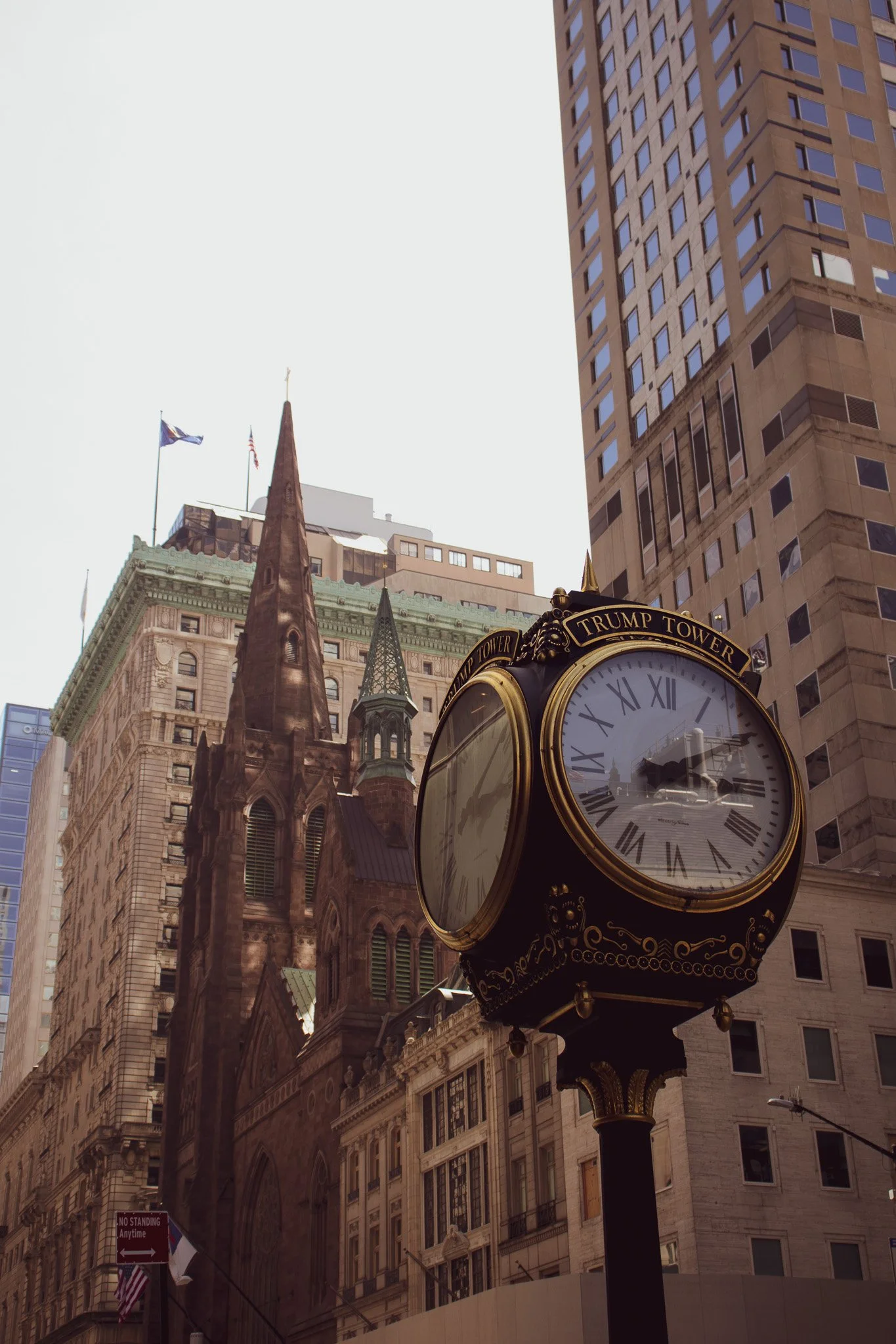 Street view showing a historical clock with 'TRUMP TOWER' inscription at the top, surrounded by high-rise buildings and a church with a steeple, American flags, and a no standing anytime sign.