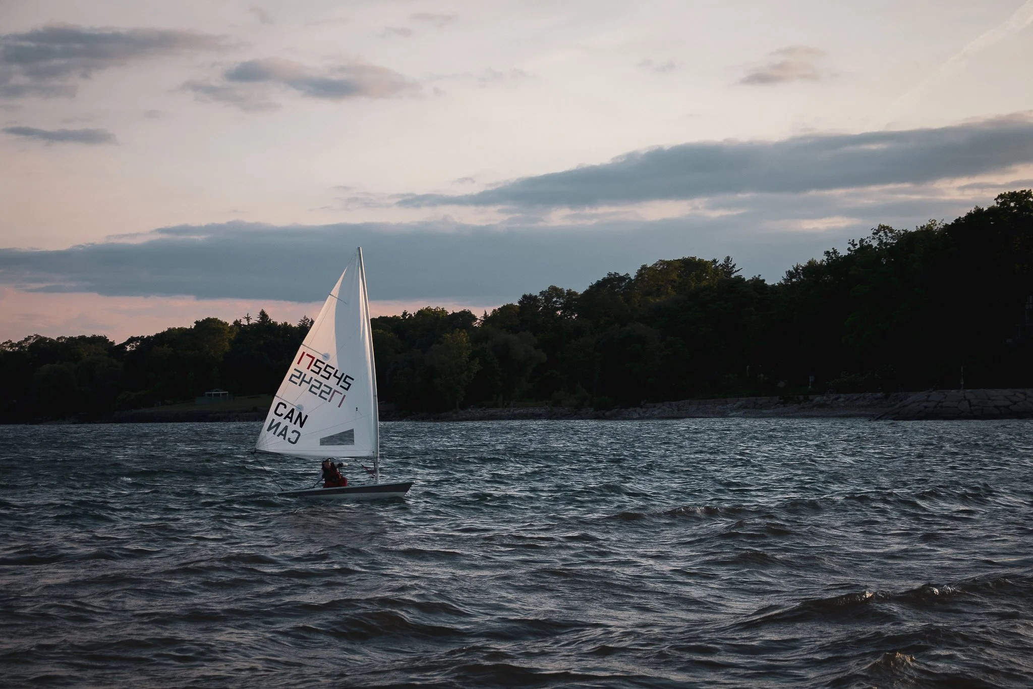 A person sailing a small boat with a white sail on a calm body of water, with a forested shoreline and a cloudy sky in the background during dusk or dawn.