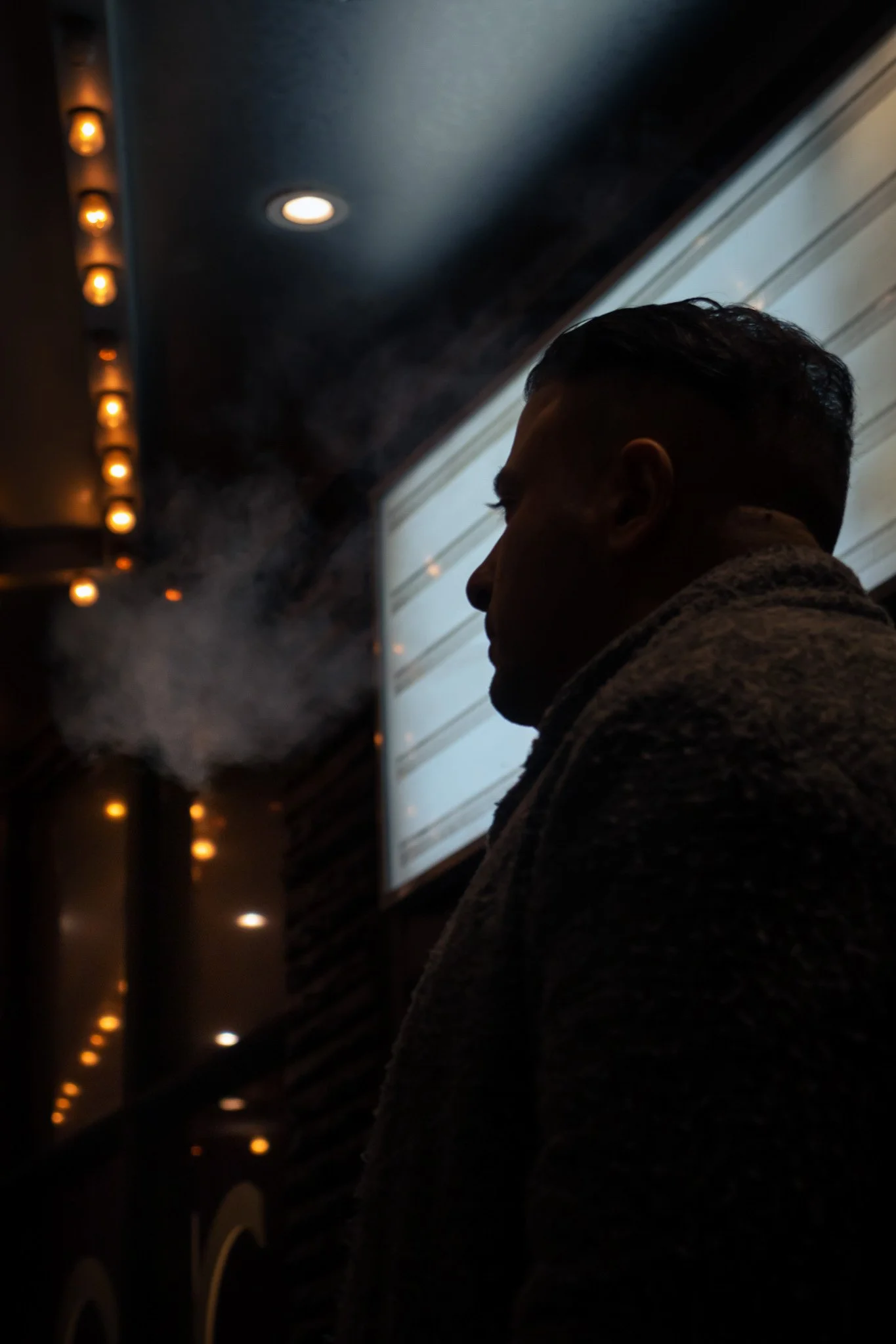 Silhouette of a person with short hair in a dimly lit indoor setting, with small warm lights along the ceiling and a bright round ceiling light.