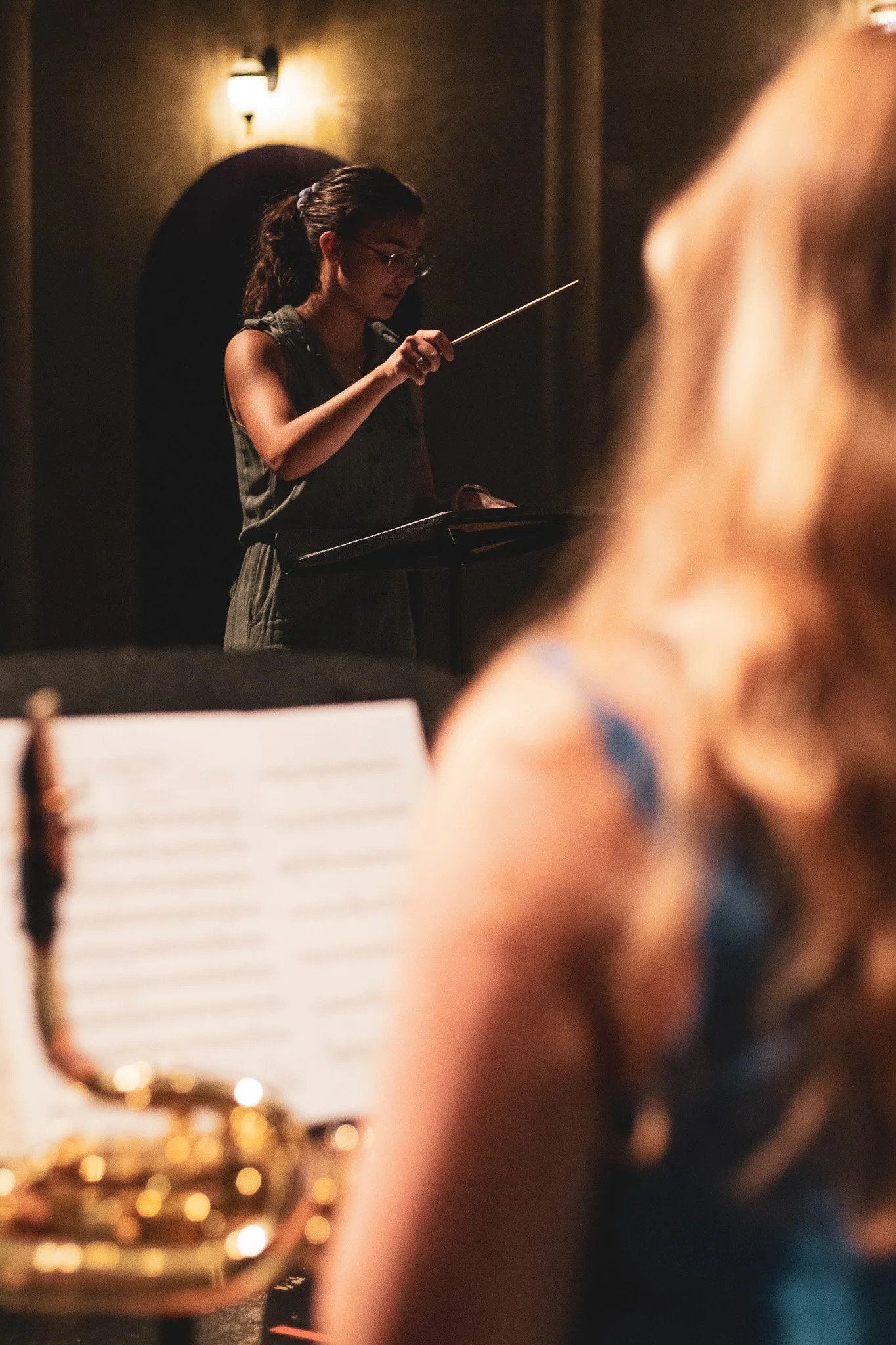 A woman with glasses and dark hair conducting an orchestra, seen through a musical instrument's bell, in a dimly lit room.