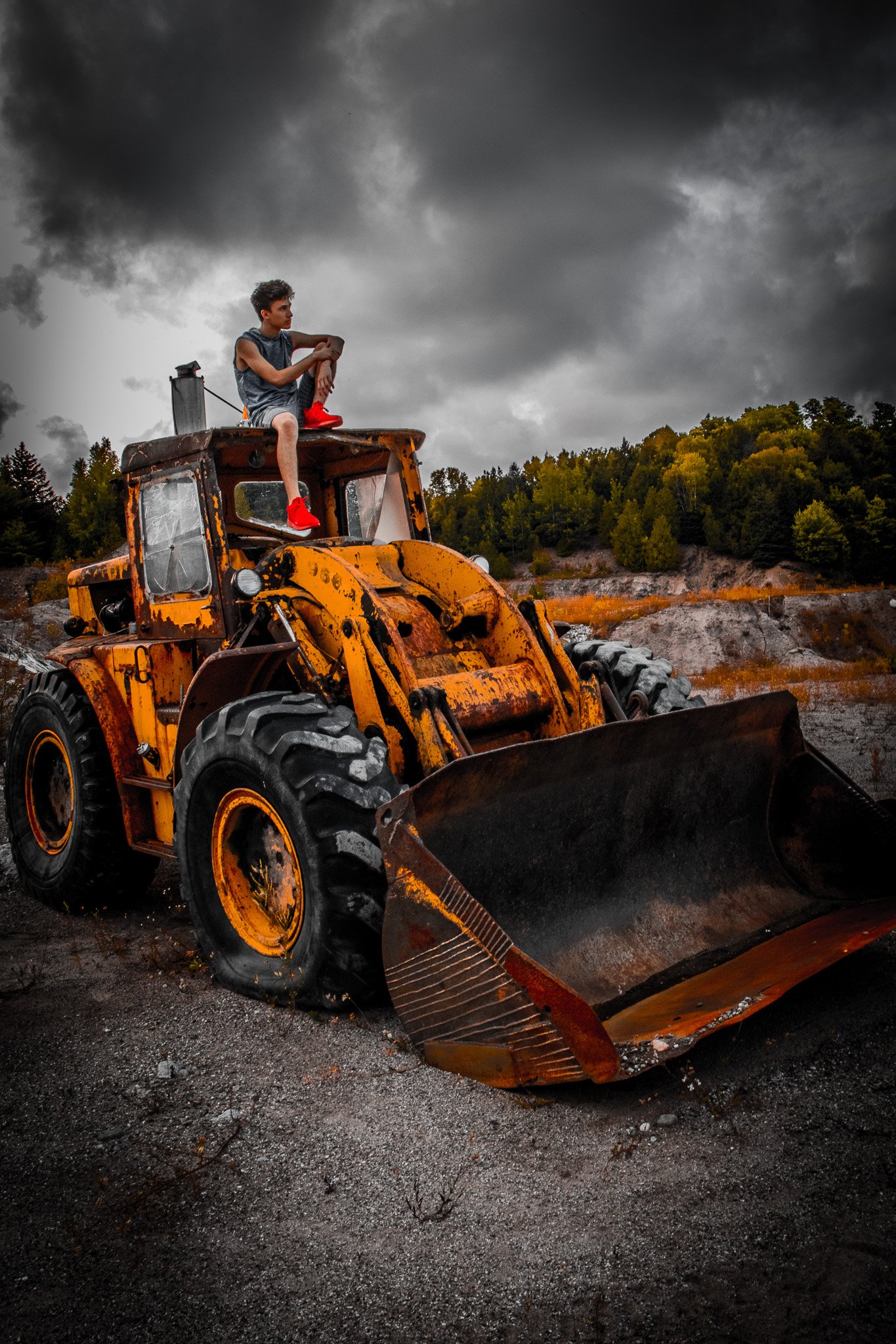 A young man sitting on top of a large, rusted, yellow front loader bulldozer, looking contemplative while holding his left arm with his right hand, on a dirt and gravel surface outdoors during a stormy day with dark clouds and a forested hillside in 