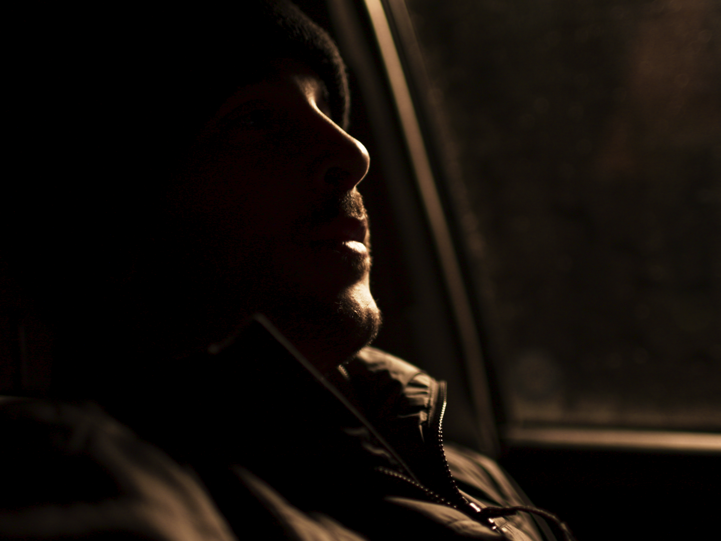 A person is sitting inside a vehicle with their eyes closed and face turned slightly upward, illuminated by a warm light from the side, possibly during sunset or sunrise.