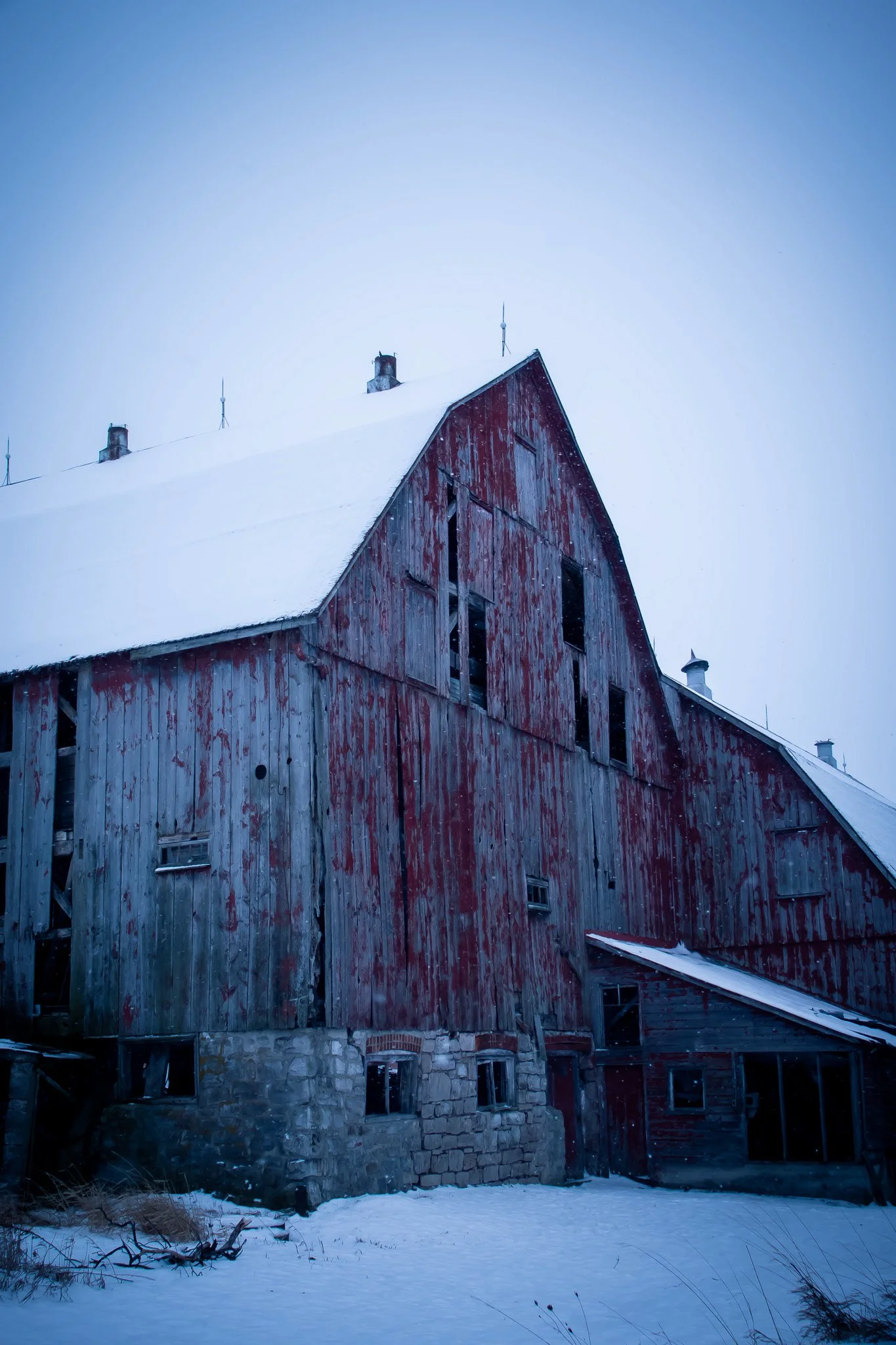 Old wooden barn with red peeling paint, snow covering the roof and ground, small windows, and chimney pipes, set against a pale sky in winter.