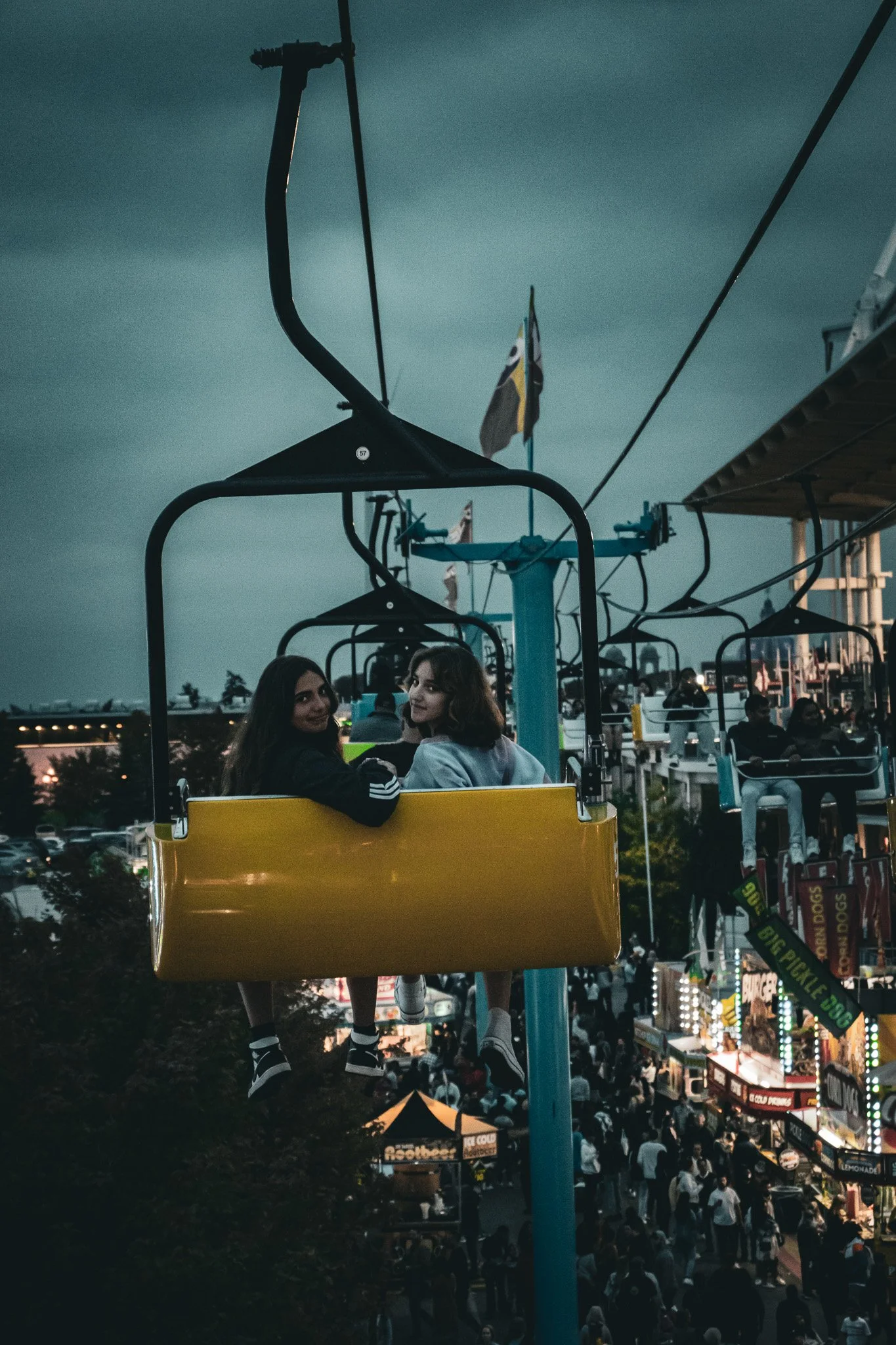 Two young women are riding a yellow gondola on a Ferris wheel at a fair or amusement park during the evening. The background shows a large crowd of people, food stands, and bright lights.