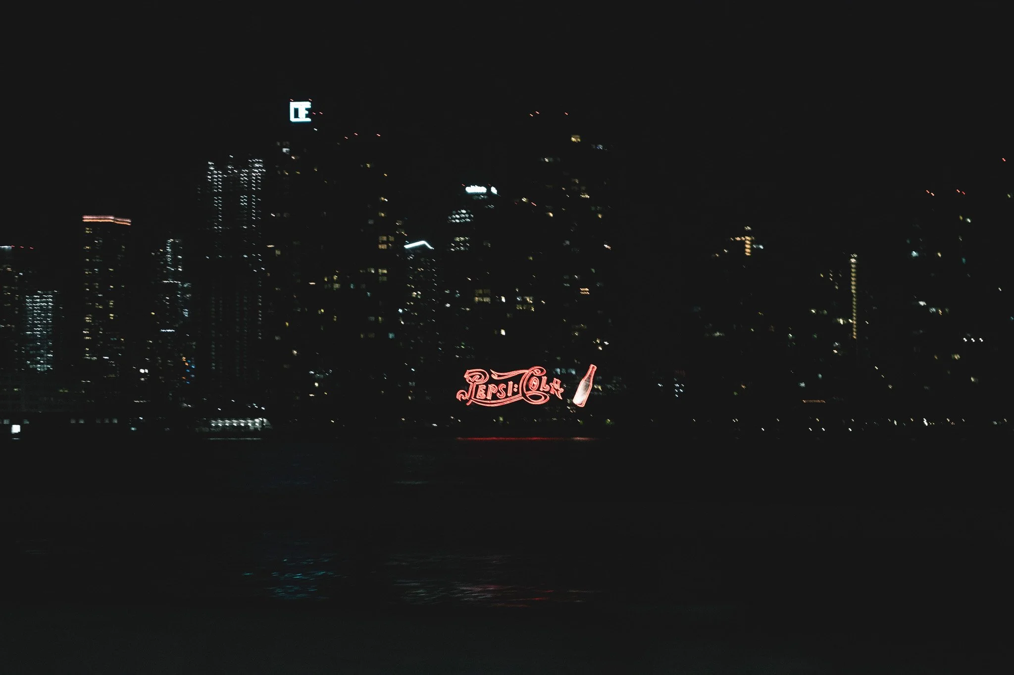 Nighttime cityscape with illuminated buildings, including a Pepsi-Cola sign and a lit-up skyscraper with a prominent logo.