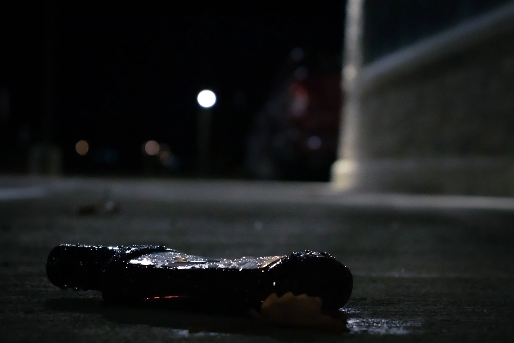 A close-up of a wet object, possibly a clump of ice or a piece of debris, on a dark surface at night, with a blurred background featuring lights and a faint outline of a building or structure.
