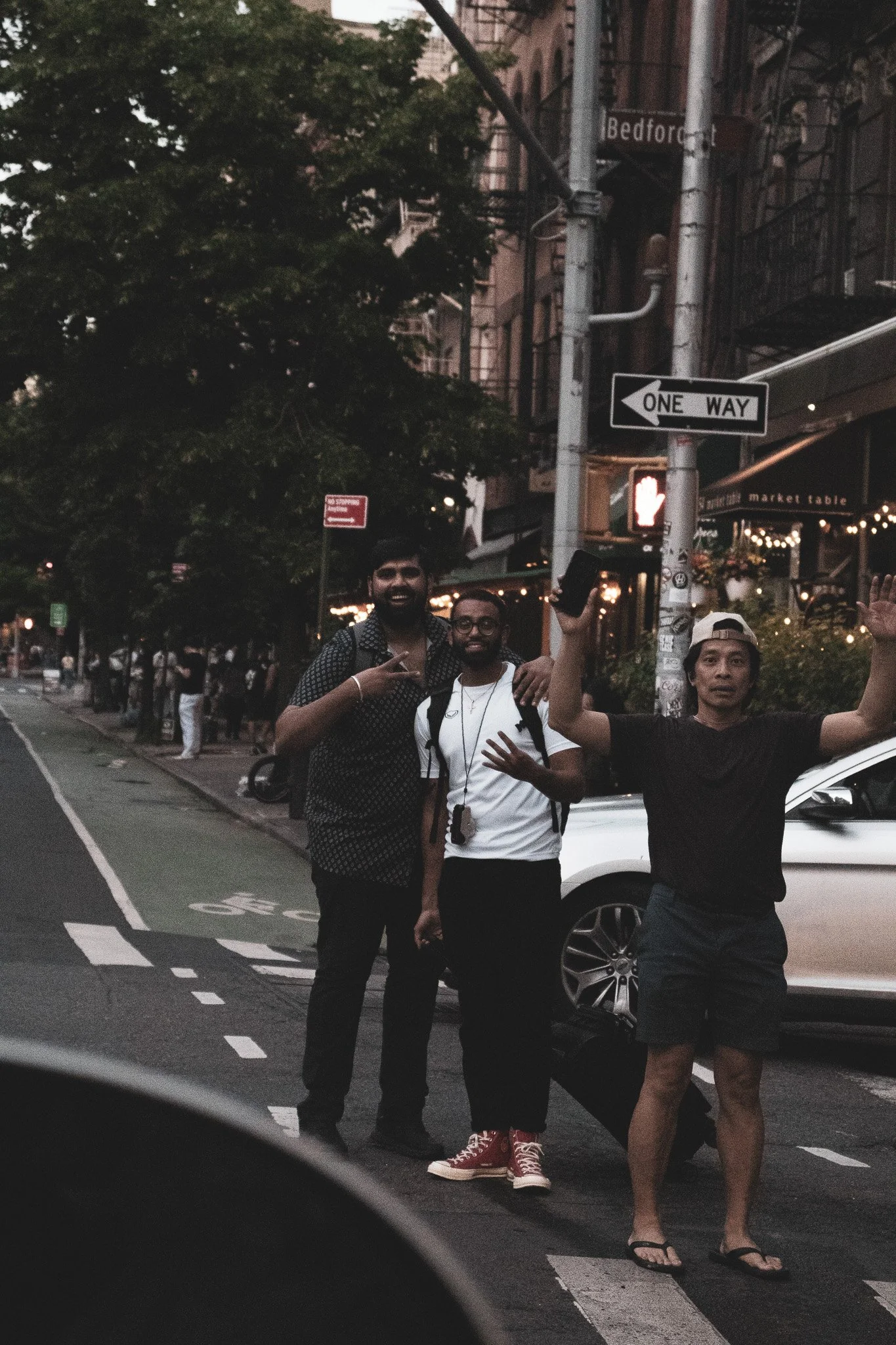Three men standing on a city street, two smiling and one with arms outstretched, near cars and buildings, at dusk.