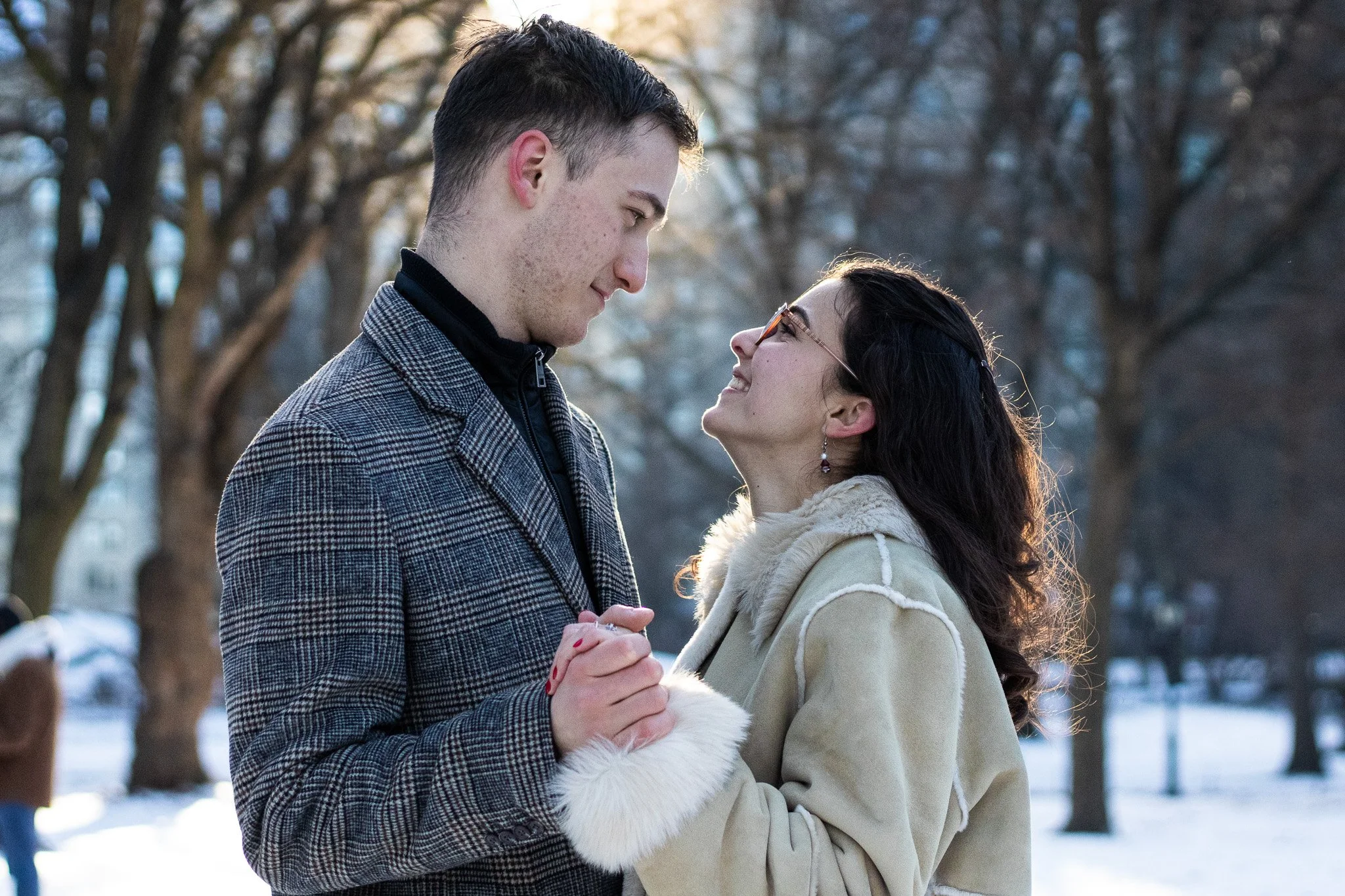 A young man and woman standing close together outdoors in winter, holding hands and gazing at each other, with leafless trees and snow on the ground in the background.