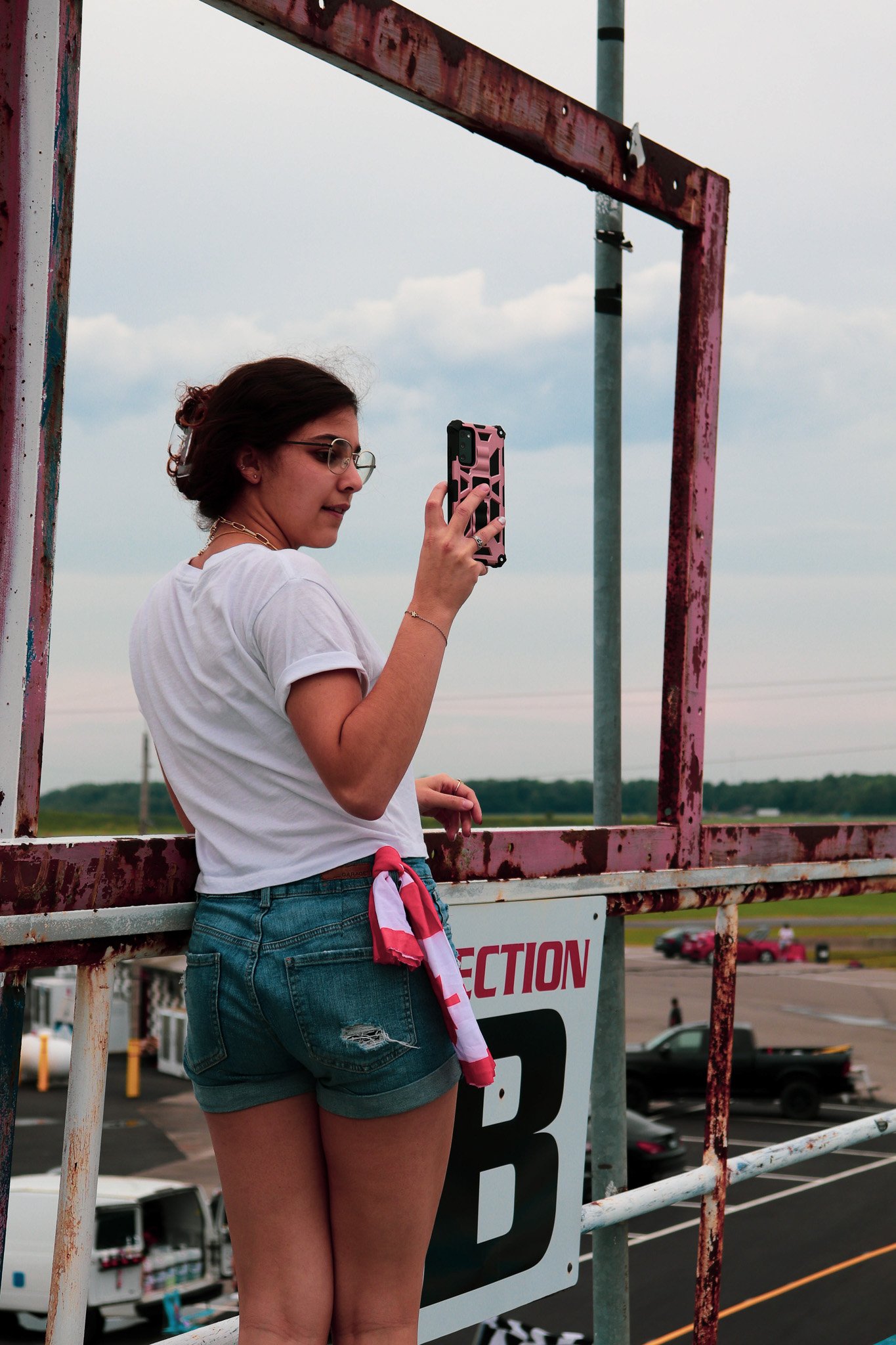 A woman with short dark hair, wearing glasses, a white t-shirt, denim shorts, and a red and white cloth tied to her waist, is taking a photo with her phone from a dirt parking lot with a sign that says 'Section B'. She stands next to rusted metal rai